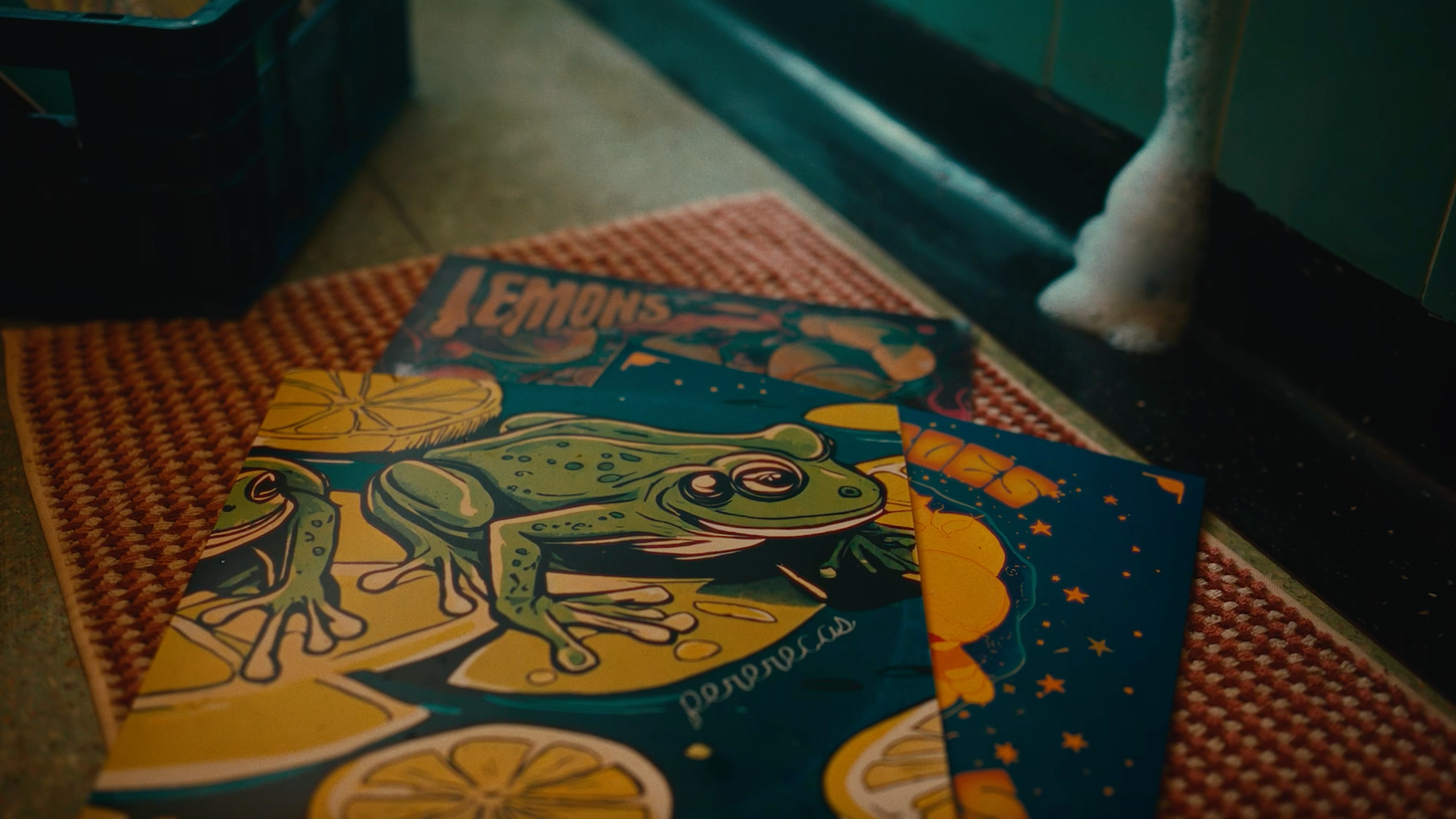 Three colorful posters with illustrations of frogs and lemons lie on a red mat. One poster is partially labeled LEMONS. A white animals paw, possibly a cat, is visible on the right edge of the image.