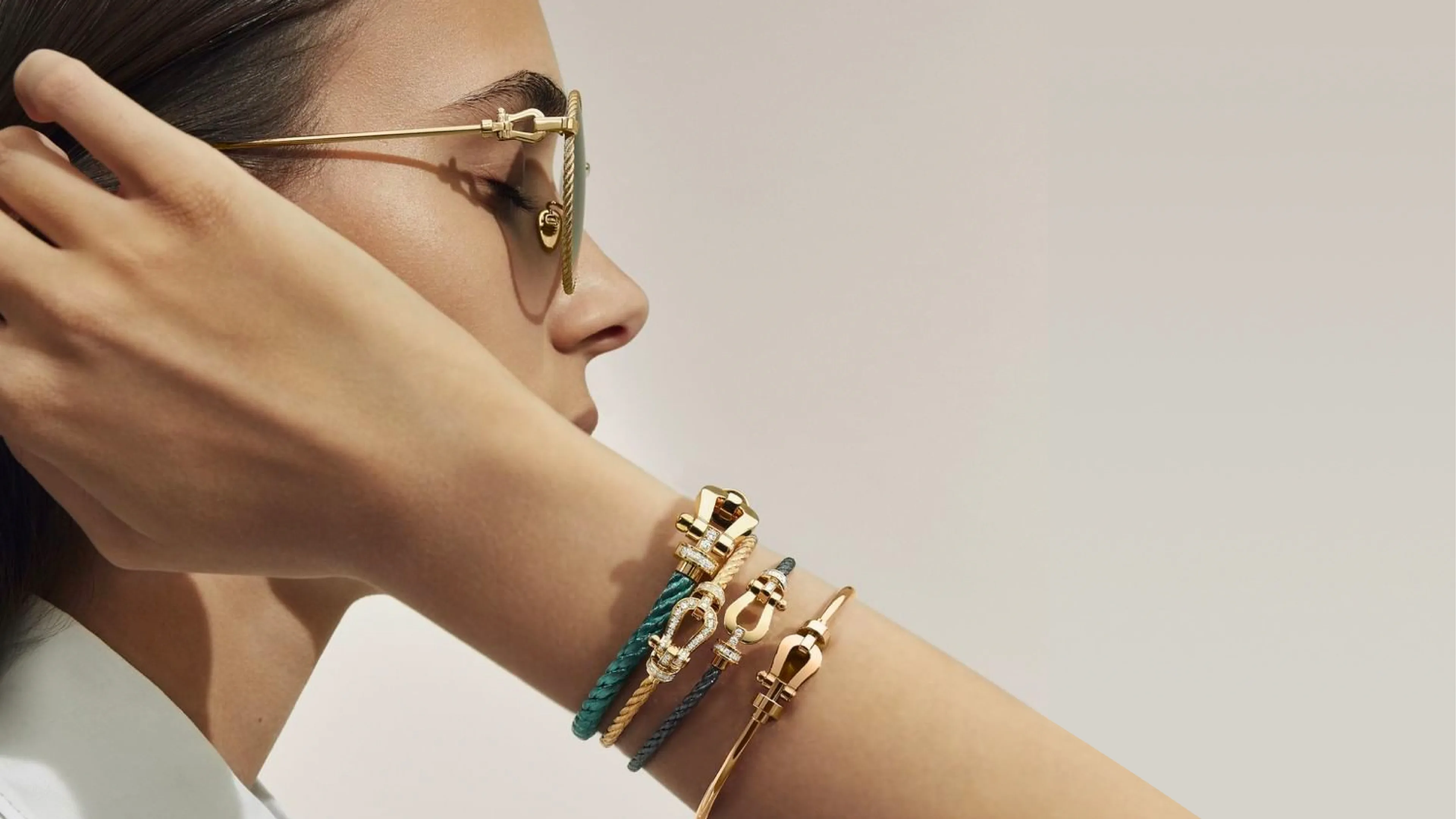 A woman in profile adjusts gold-rimmed sunglasses; her arm is raised, displaying several gold and blue bracelets against a neutral background.