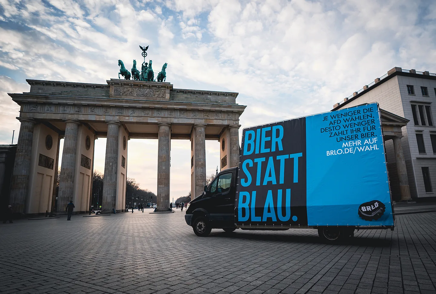 A truck with a large sign reading “Bier statt Blau.” is parked in front of the Brandenburg Gate in Berlin. The sky is partly cloudy, and the plaza appears mostly empty.