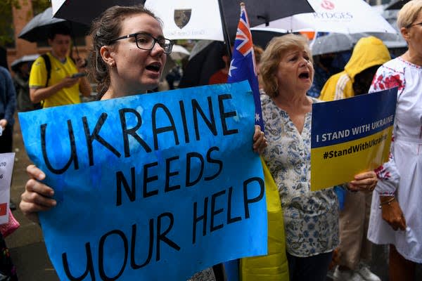 A woman holds a blue sign reading UKRAINE NEEDS YOUR HELP at a rally. Other protesters nearby hold umbrellas and signs supporting Ukraine, including one that says #StandWithUkraine.