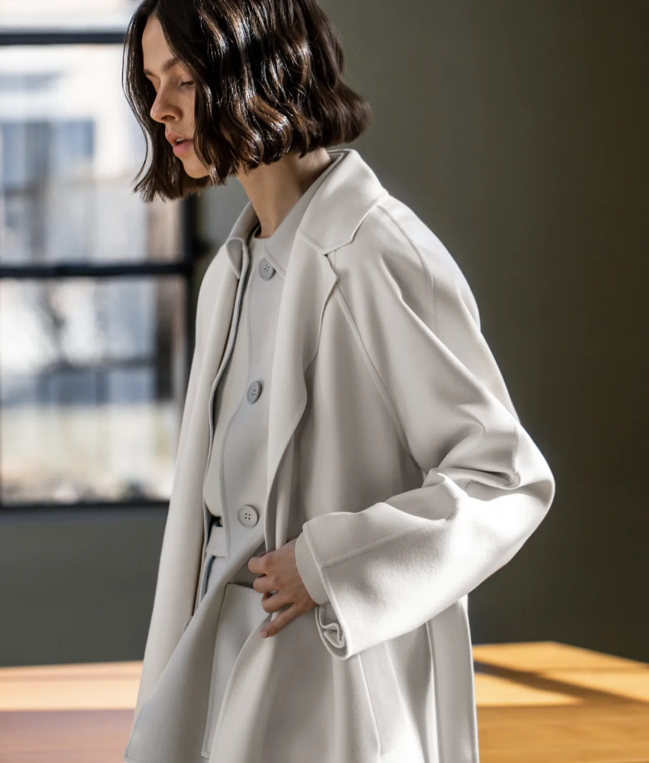 A woman with wavy, shoulder-length dark hair wearing a light-colored coat stands indoors near a sunlit window and wooden table, looking down thoughtfully.