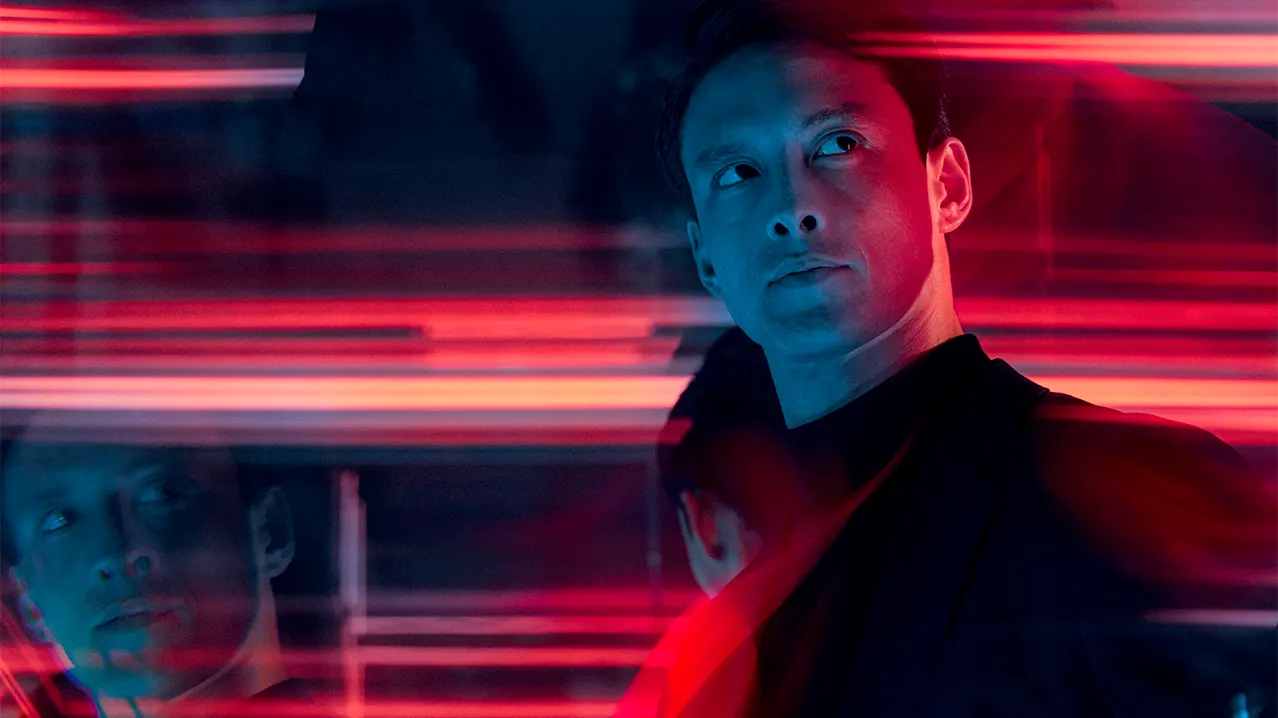 A man in a dark suit stands in moody lighting, looking to the side with a serious expression. Red and blue light streaks blur across the image, creating a dynamic, futuristic atmosphere. His reflection appears beside him.
