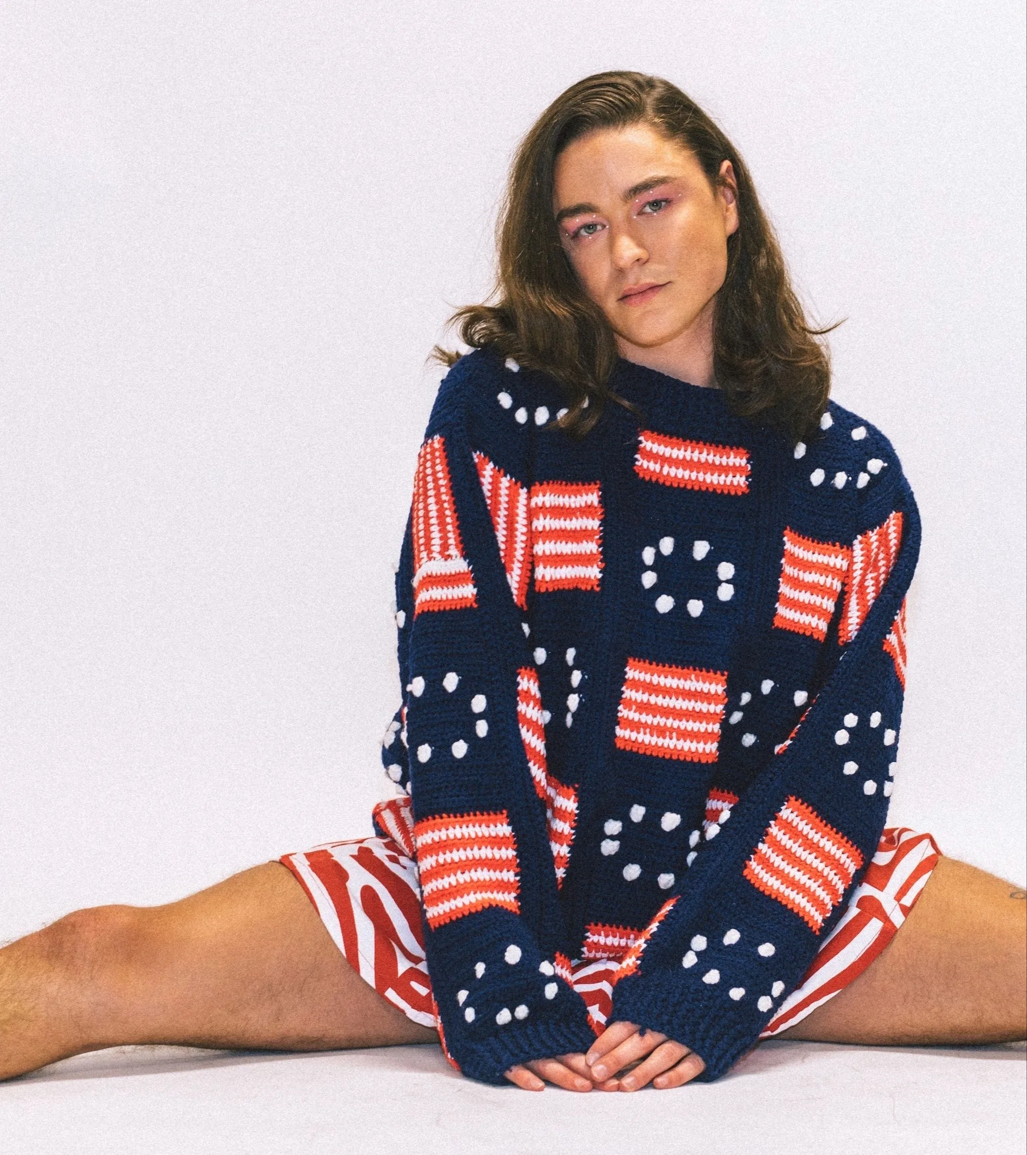 A person with long brown hair sits on the floor with legs apart, wearing a navy blue sweater with red, white, and blue geometric patterns and red-and-white striped shorts, against a plain white background.