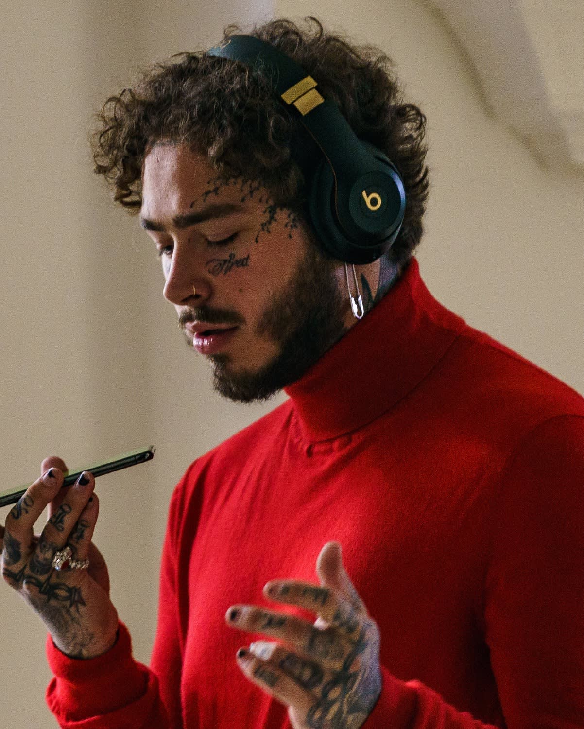 A man with curly hair, tattoos, and facial piercings wears black Beats headphones and a red turtleneck. He holds a phone in one hand and gestures with the other, appearing focused or engaged with the device.
