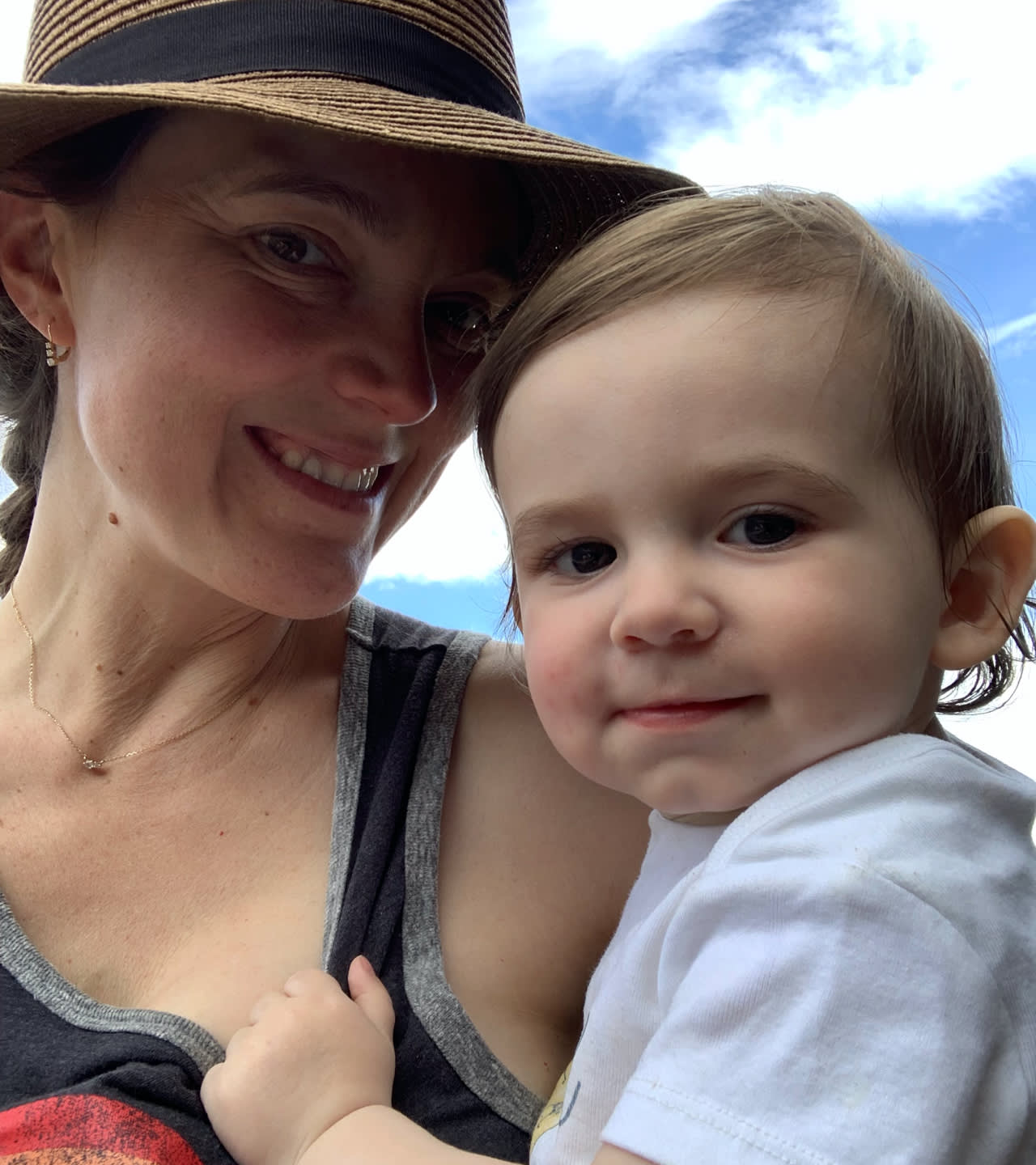 A woman wearing a hat smiles while holding a young child with short brown hair. The child, dressed in a white shirt, looks at the camera. The background shows a blue sky with scattered clouds.