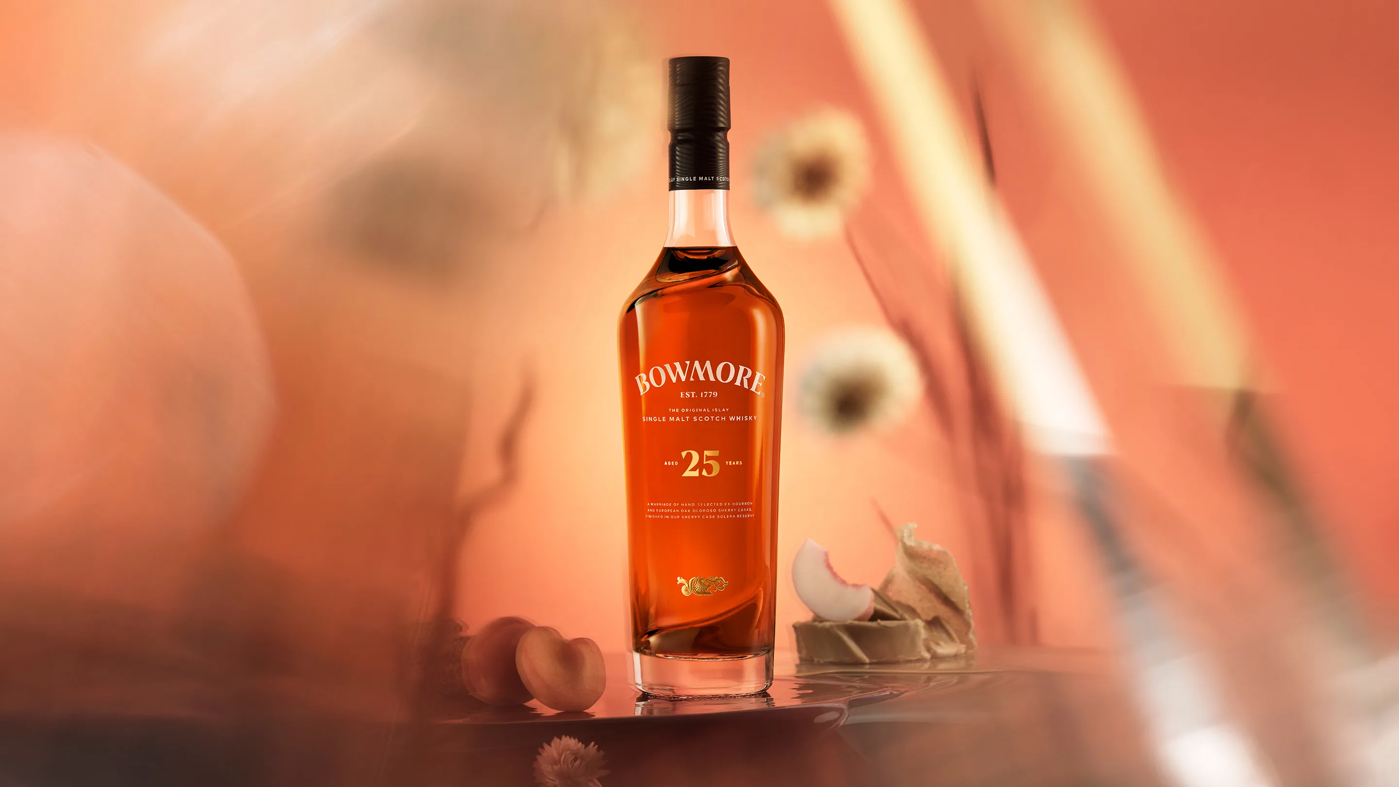 A bottle of Bowmore 25-year-old single malt scotch whisky stands on a reflective surface with a soft orange background, accompanied by dried flowers and peach slices.