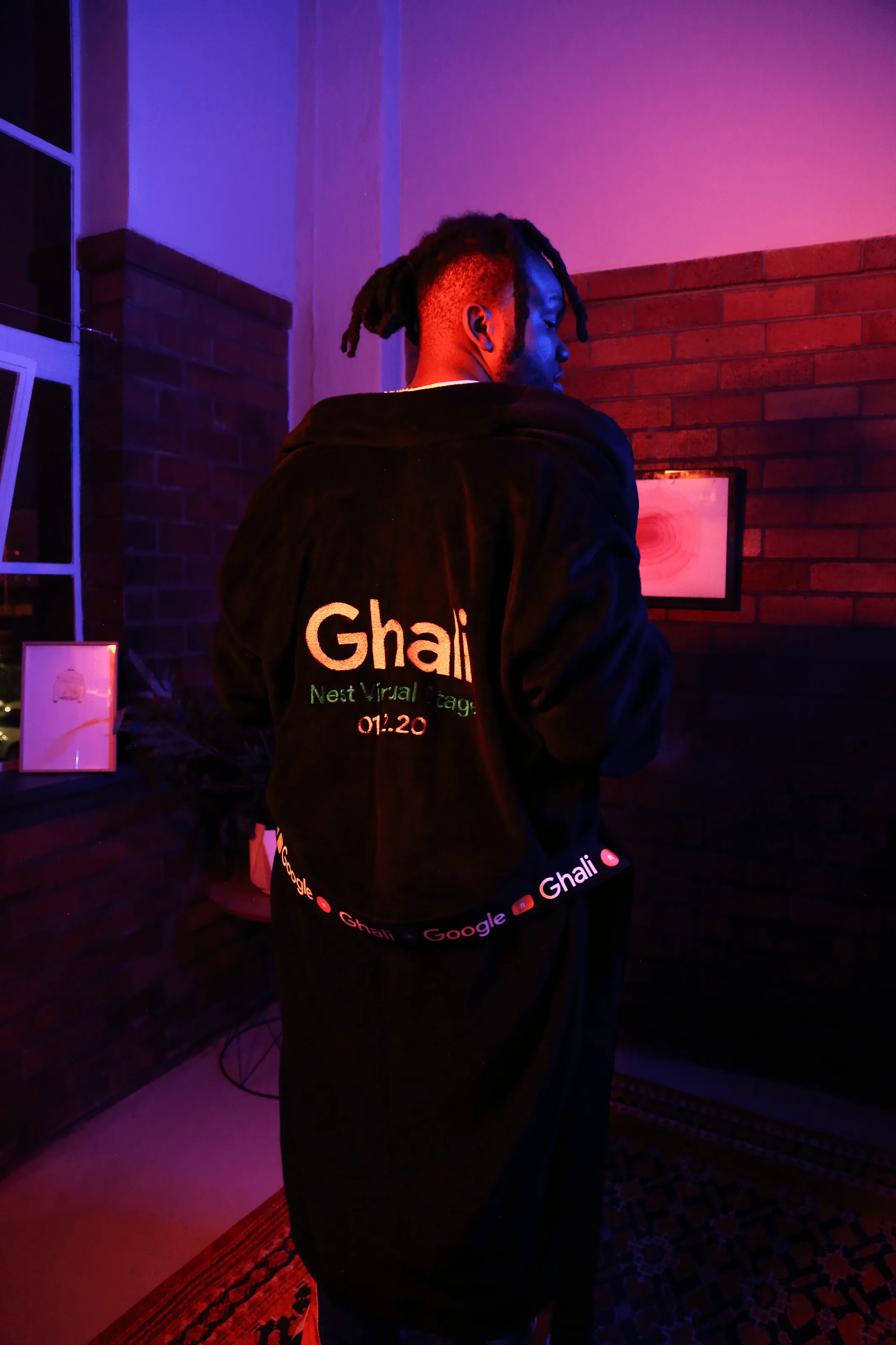 A man stands indoors at night wearing a black robe with Ghali and text on the back, facing away from the camera. The room is lit with purple and blue lights and has brick walls and a window.