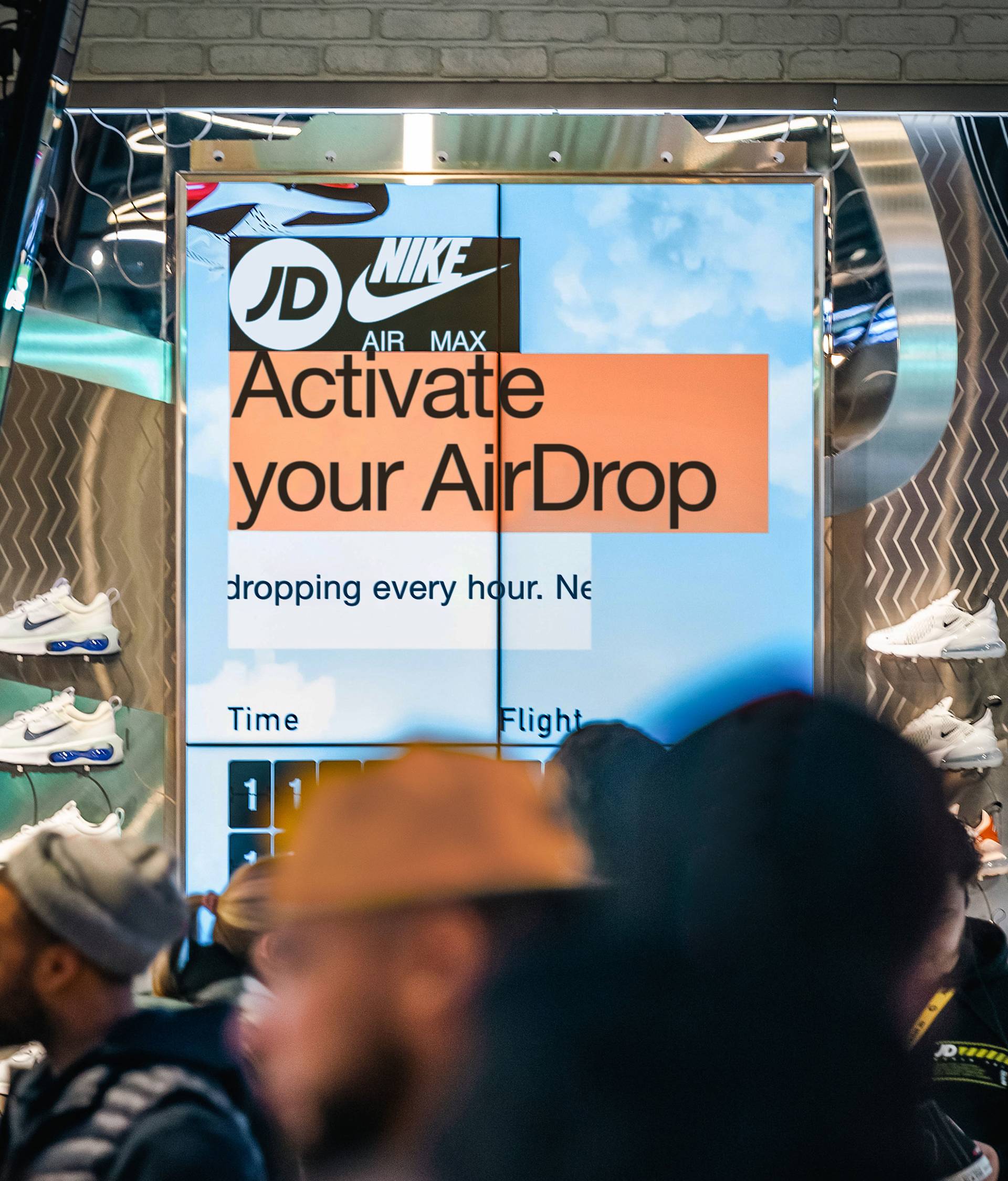 A digital sign in a shoe store displays “Activate your AirDrop” with JD and Nike logos. People in the foreground browse, and shelves with sneakers are visible in the background.