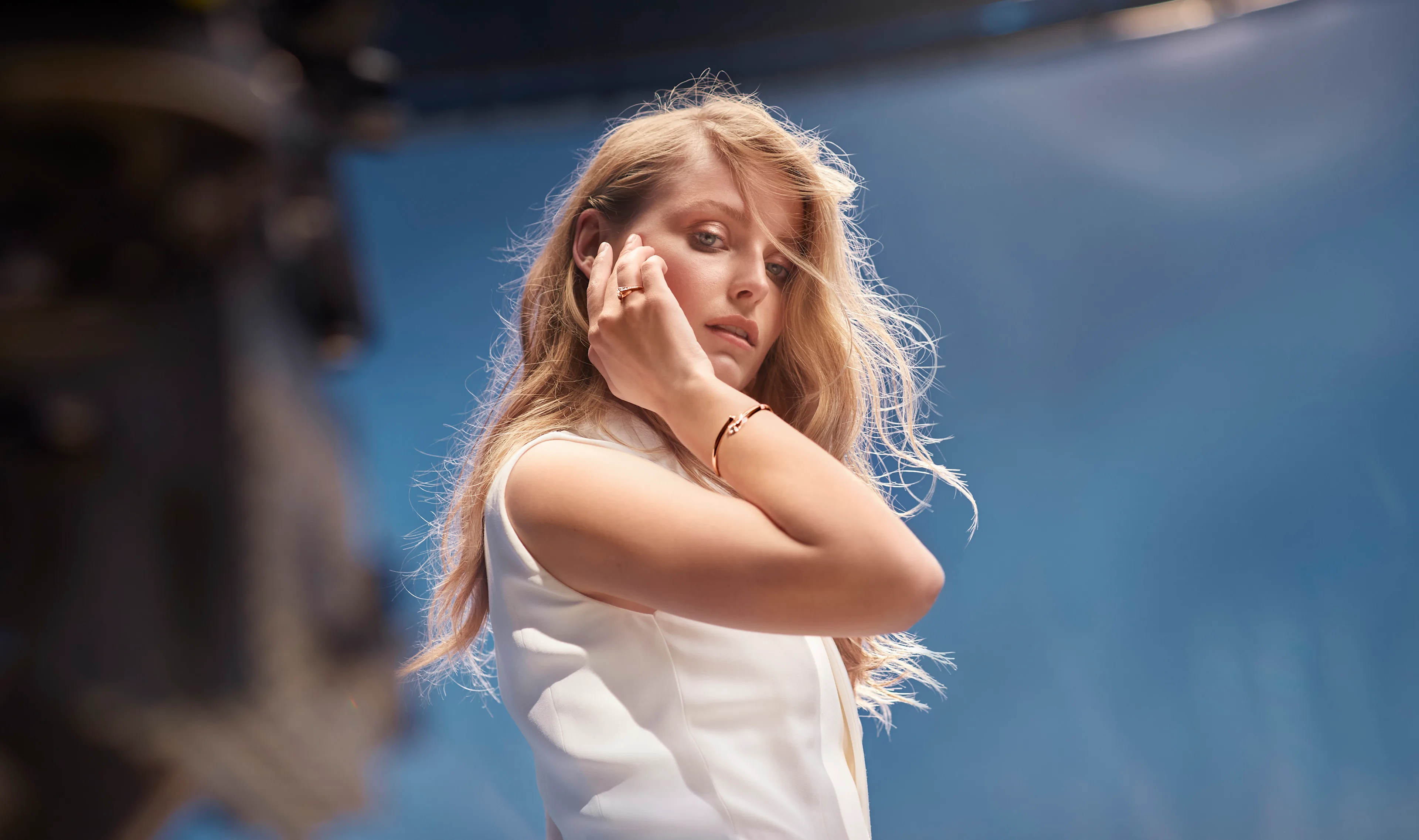 A woman with long, wavy blonde hair wearing a sleeveless white top touches her ear and looks slightly downward against a soft blue background. Light shines on her hair, creating a gentle, serene atmosphere.