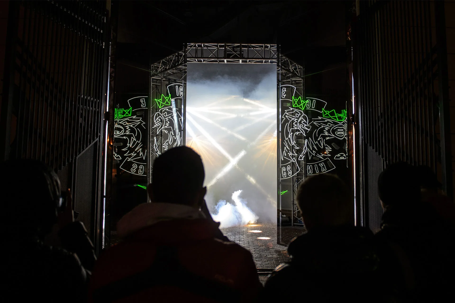 Silhouetted people face a misty, illuminated entrance with bright spotlights and artistic black panels featuring graffiti-style drawings on each side. The scene has a dramatic, futuristic atmosphere.