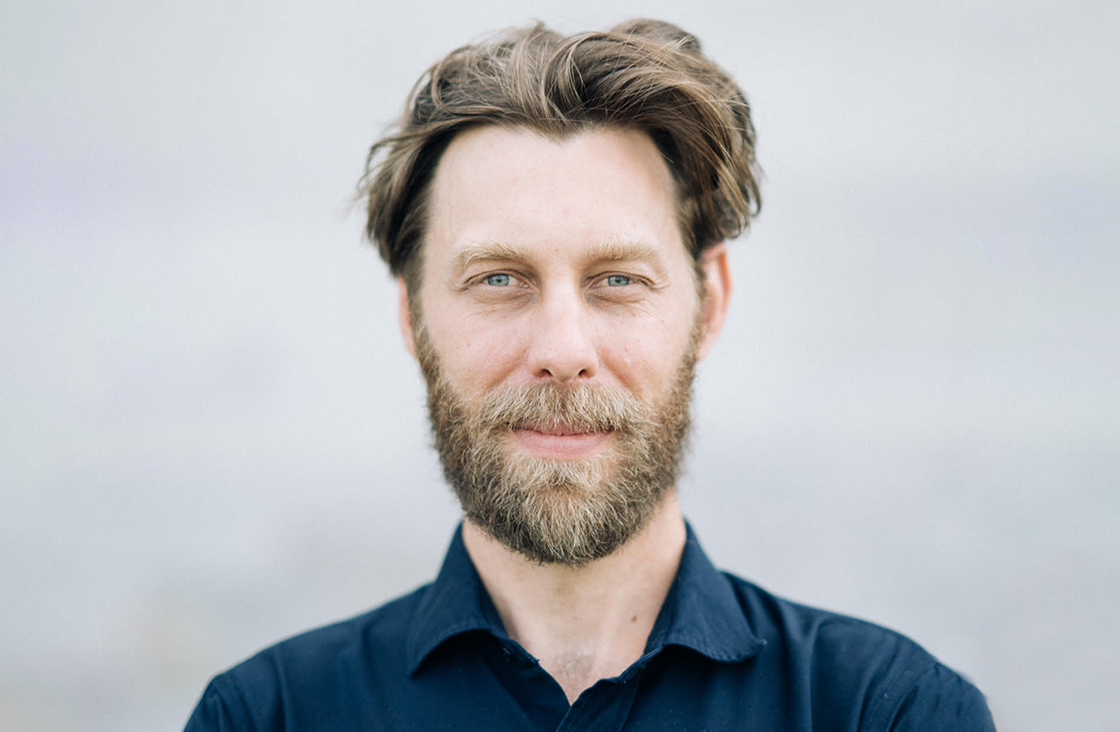 A man with light skin, blue eyes, and a full brown beard stands outdoors, looking at the camera. He has medium-length brown hair and is wearing a dark shirt. The background is blurred and light-colored.