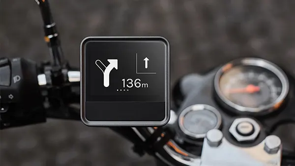 A digital navigation device mounted on a motorcycle handlebar displays a turn-by-turn direction, showing a right turn in 136 meters. The motorcycle speedometer is partially visible in the background.