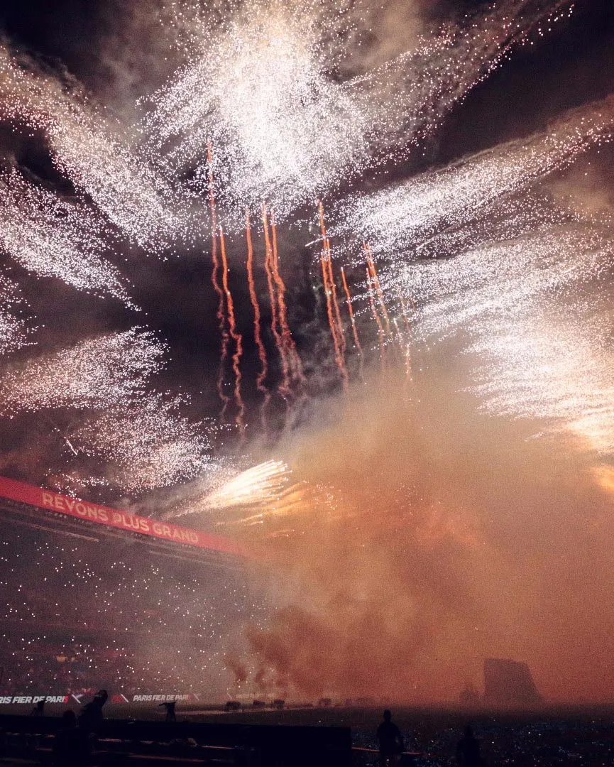 Bright fireworks burst in the night sky above a stadium, filling the scene with sparkling light and smoke. A red LED sign reads “RÊVONS PLUS GRAND,” and the stands are crowded with spectators.