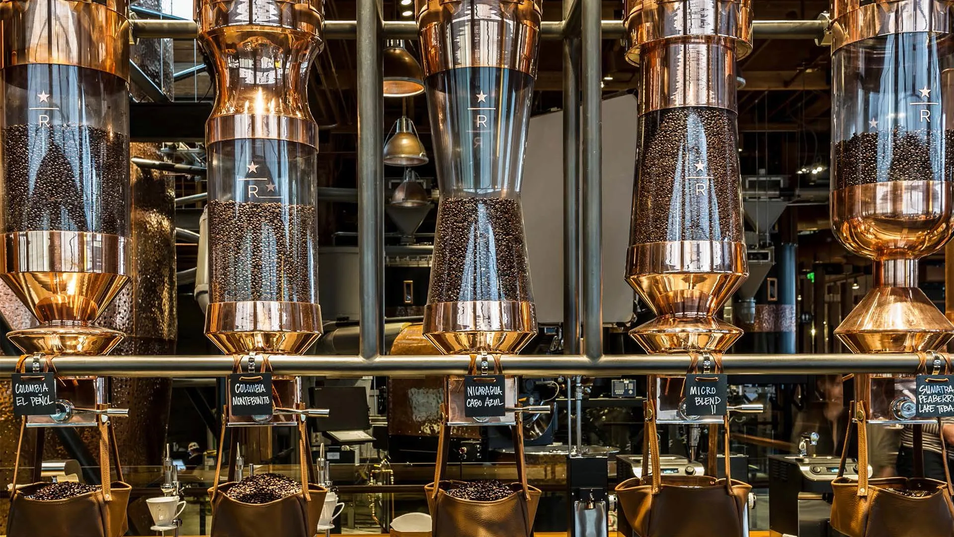 Large transparent coffee bean dispensers with copper accents are lined up above a counter in a modern coffee shop or roastery, filled with different types of coffee beans labeled below each container.