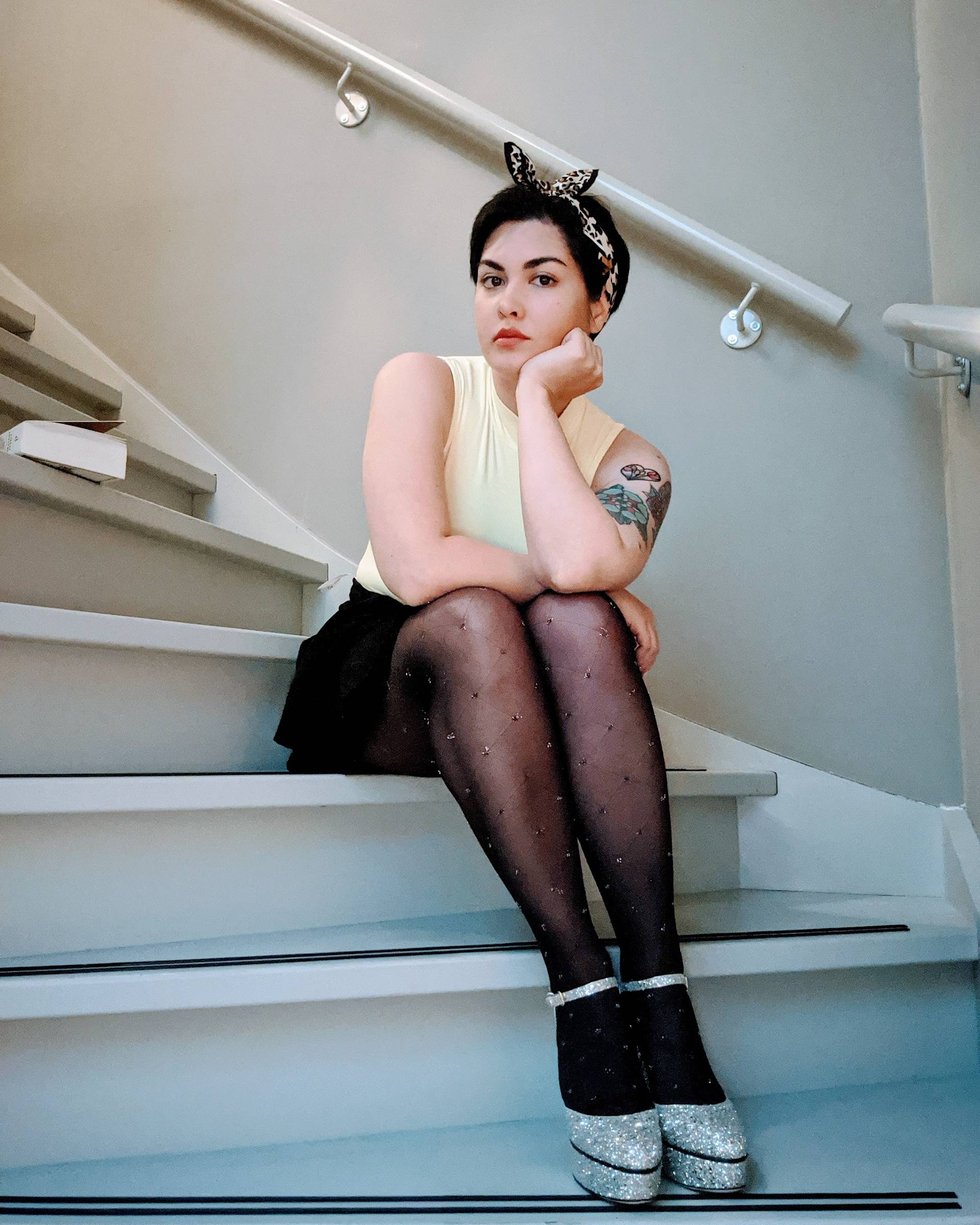 A person with short dark hair, wearing a headband with ears, a sleeveless yellow top, black skirt, sheer black tights, and glittery heels, sits on stairs with one hand resting on their chin and legs crossed.