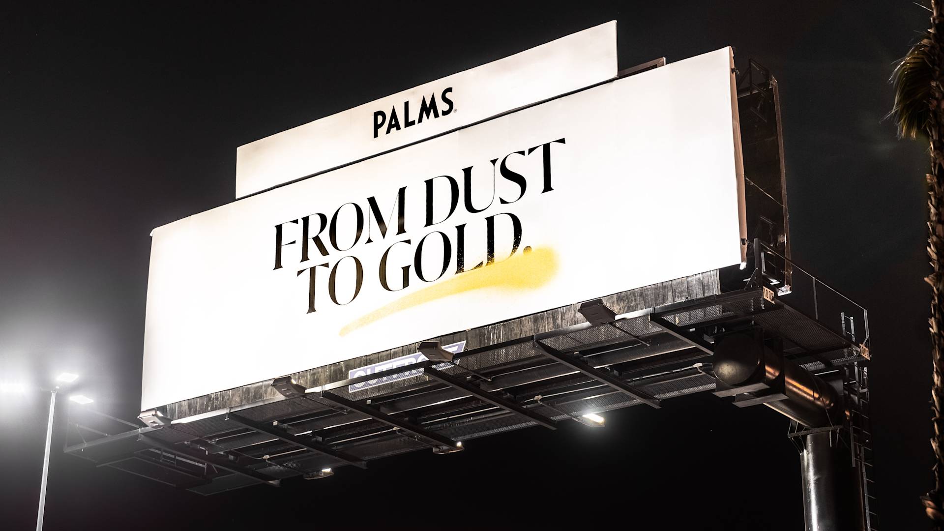 Large outdoor billboard at night reads PALMS. FROM DUST TO GOLD. with a gold brushstroke under the text. The billboard is brightly lit against a dark sky.