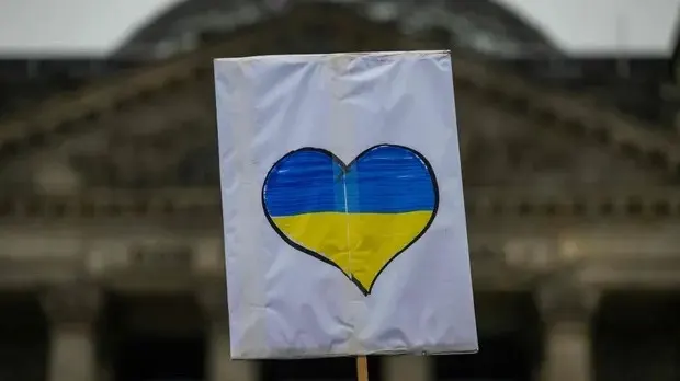 A sign with a heart-shaped design in blue and yellow, resembling the Ukrainian flag, held up in front of a blurred building.
