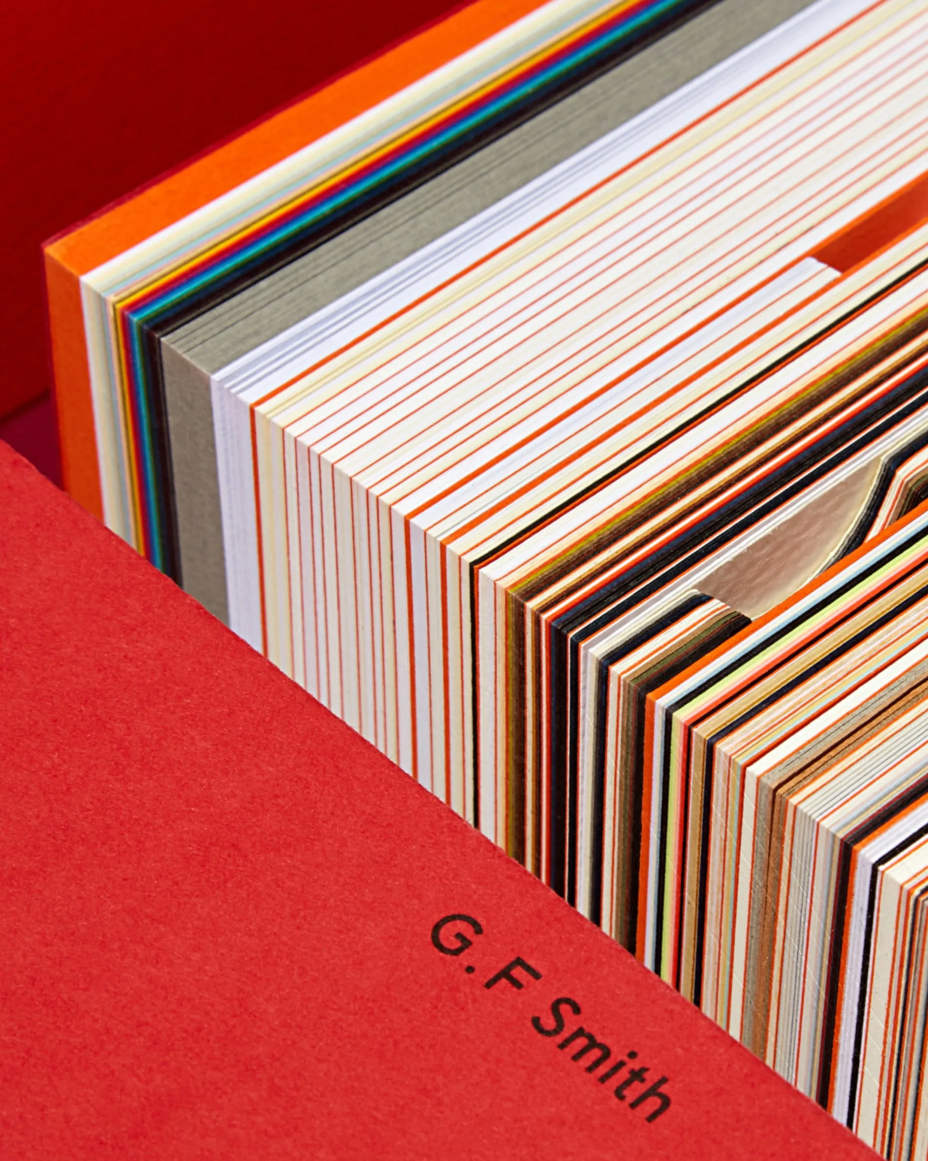 A close-up of a stack of thick, colorful paper samples with exposed edges, arranged in a red folder labeled G. F Smith. The papers are neatly layered, showcasing various colors and textures.