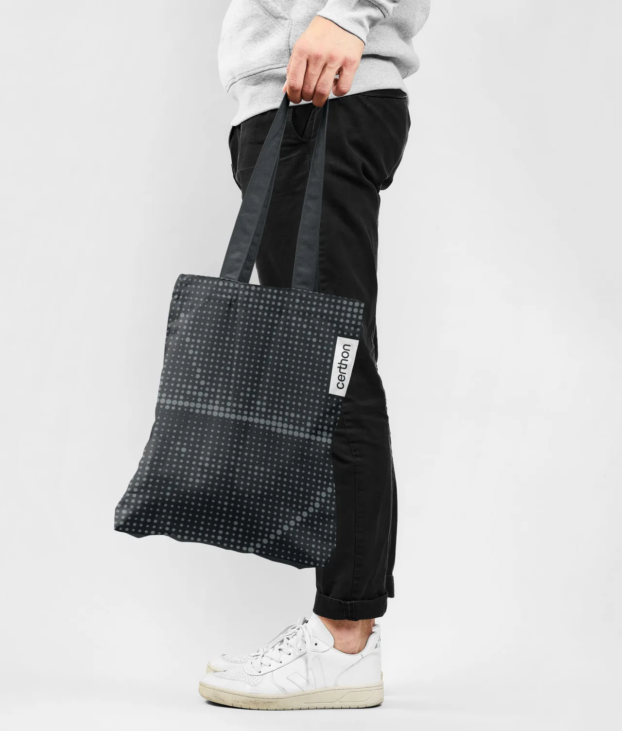 A person wearing black pants, white sneakers, and a light gray sweatshirt holds a black tote bag with a white label and small white dot pattern against a plain light background.