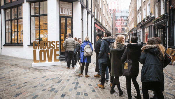 People stand in line on a cobblestone street outside a shop with large gold letters spelling CHOOSE LOVE in the window. The area appears to be a shopping district with brick buildings and window displays.