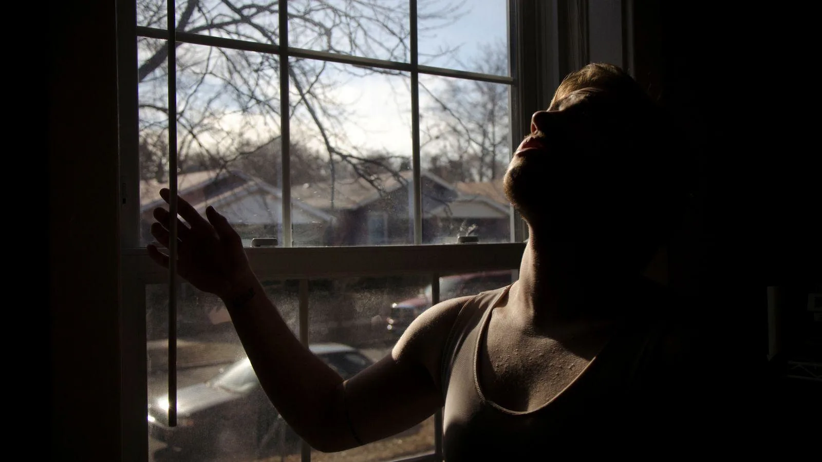 A person in a tank top stands by a window, looking upward as sunlight and shadows partially illuminate their face and body. Bare tree branches and houses are visible outside.