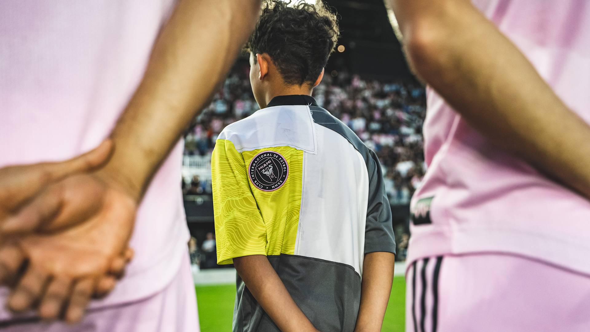 A young person in a multicolored jersey with an Inter Miami CF patch stands on a soccer field, facing away, between two players in pink uniforms with their hands behind their backs. A blurred crowd is visible in the background.