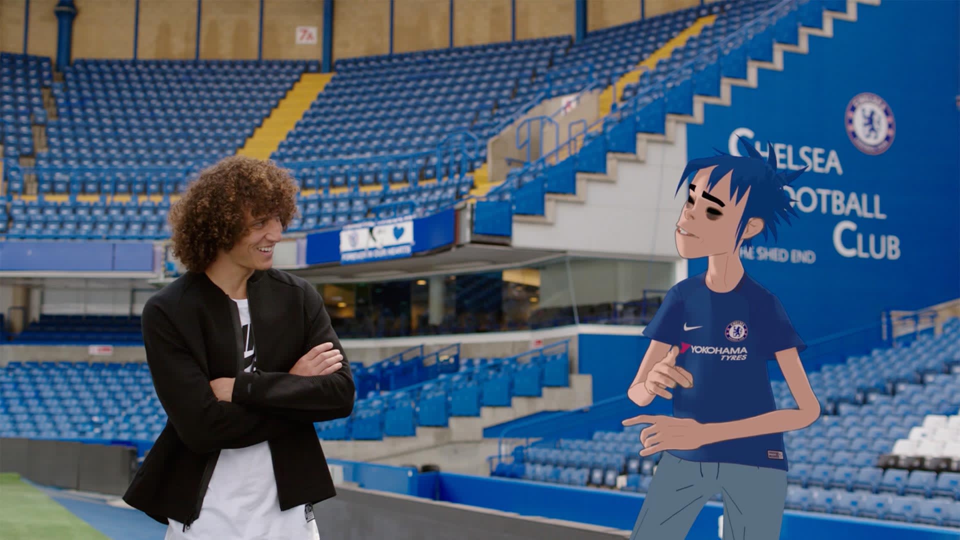 A man with curly hair, wearing a black jacket and white pants, stands smiling with arms crossed in a Chelsea FC stadium next to an animated character in a Chelsea shirt. The stands and club logo are visible in the background.