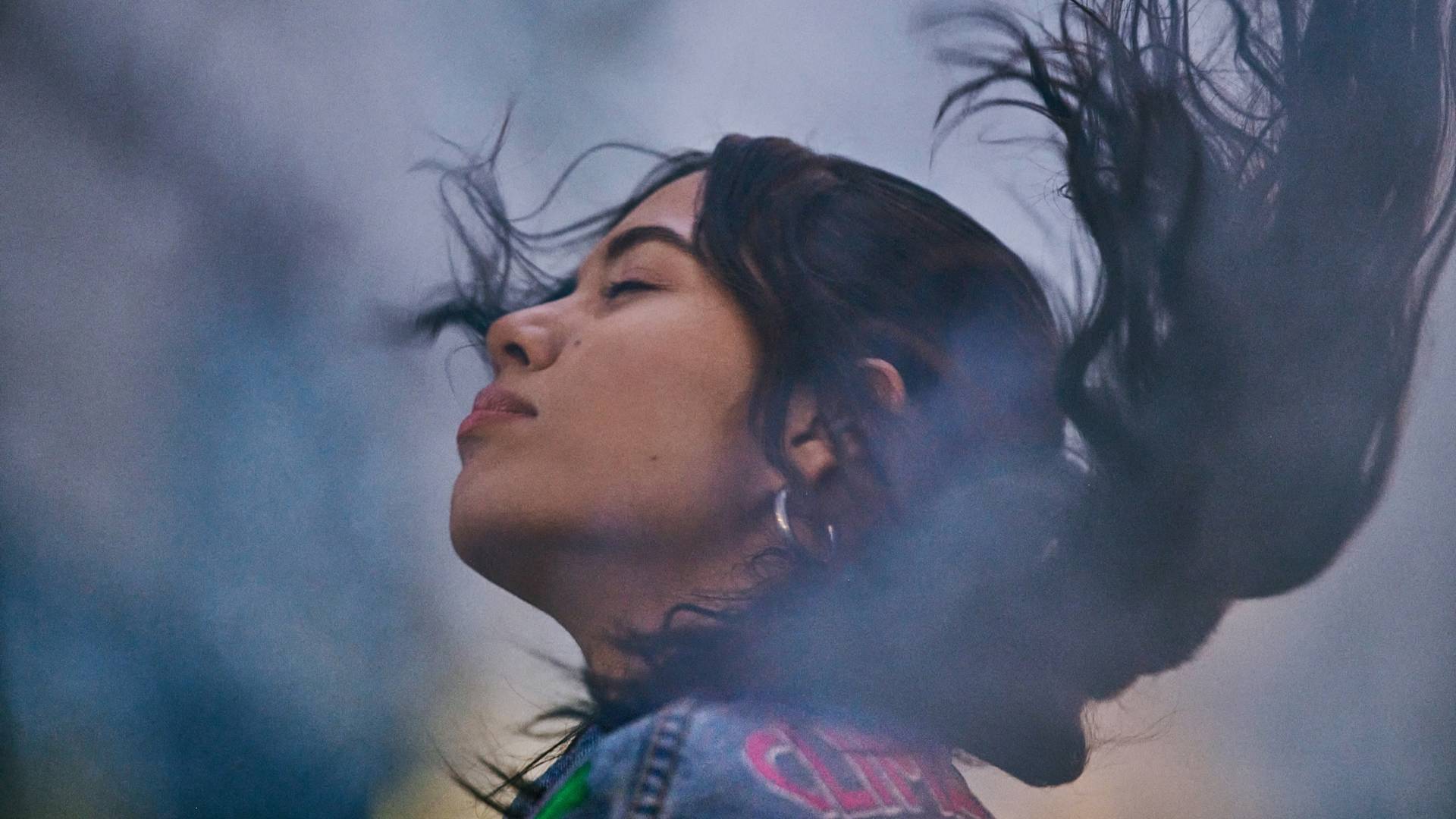 A young woman with long dark hair closes her eyes and gently tilts her head back, hair swirling in motion, creating a dreamy and serene atmosphere with soft, blurred colors in the background.