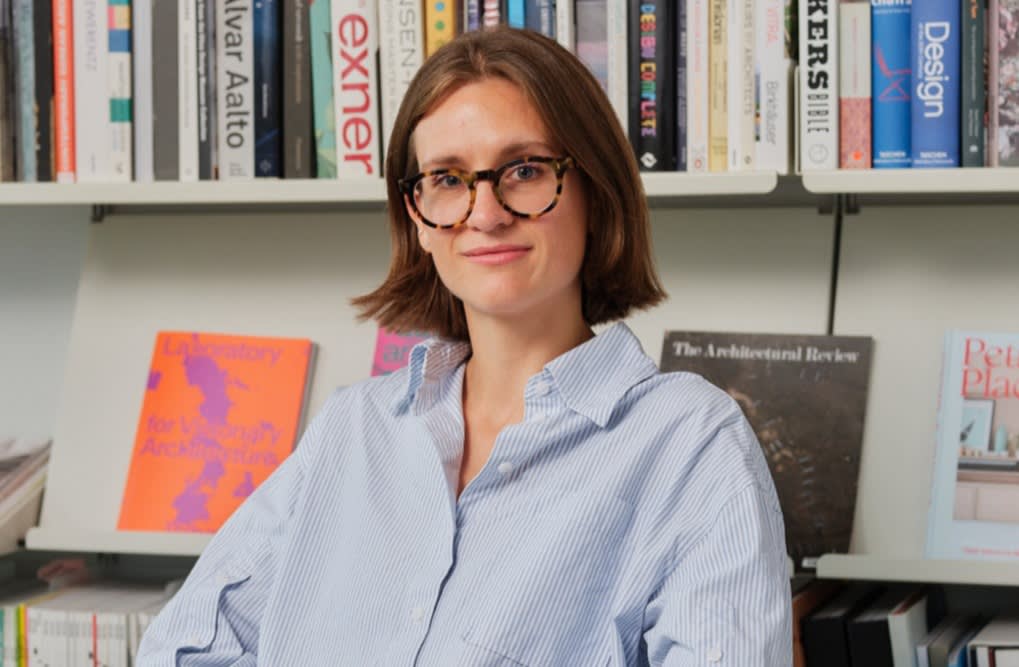 A person with shoulder-length brown hair and glasses sits in front of bookshelves filled with books and magazines, wearing a light blue button-up shirt and smiling slightly at the camera.