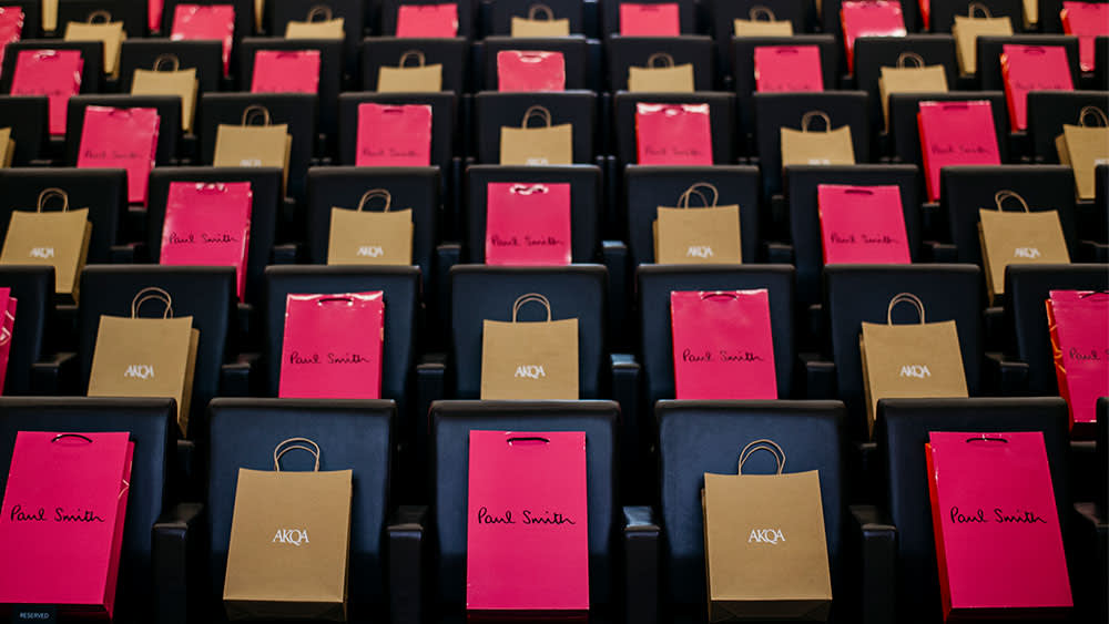 Rows of black seats with alternating pink Paul Smith and brown AKQA gift bags placed on each seat, creating a colorful, organized pattern in a large auditorium or event space.