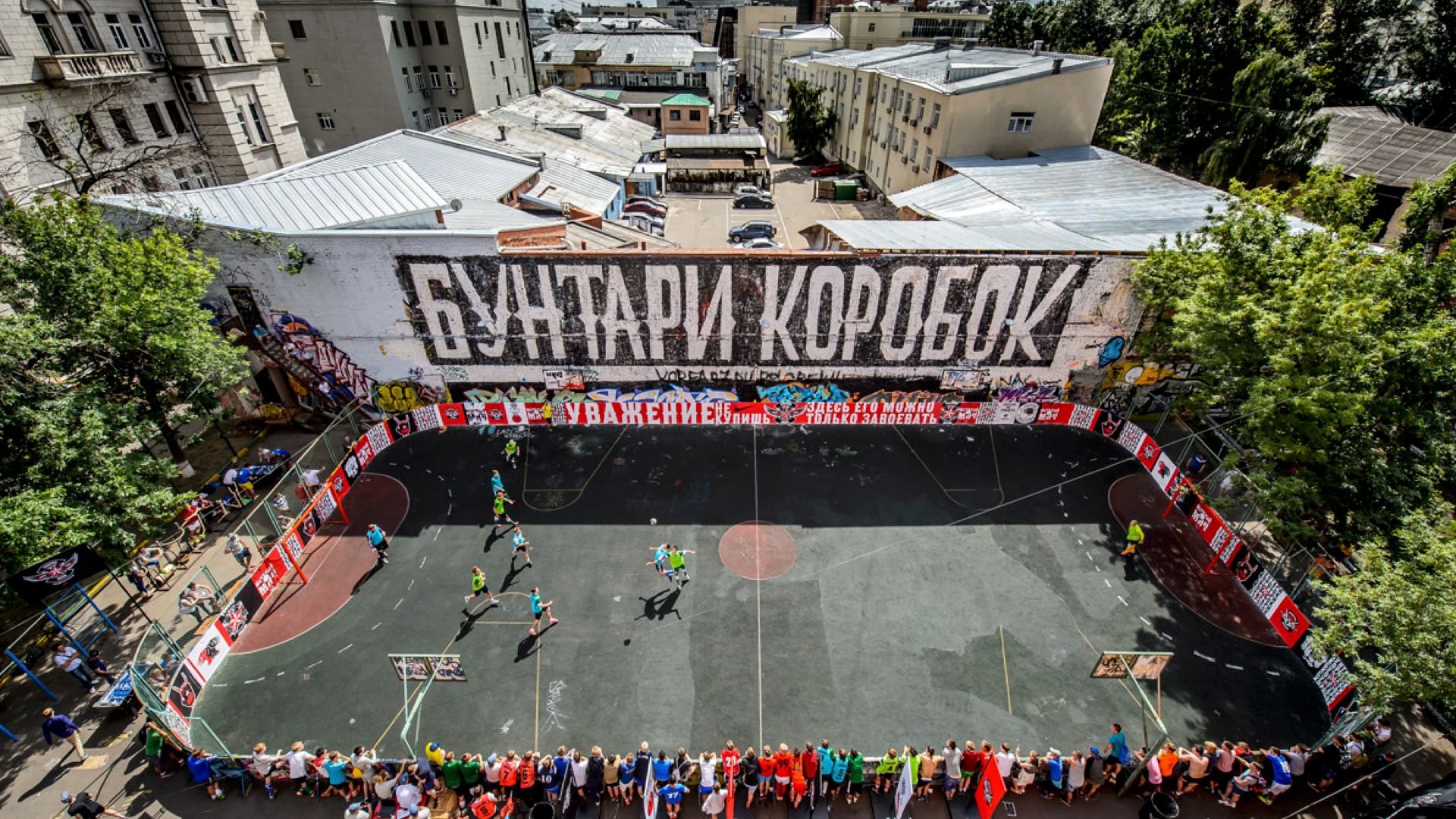 Aerial view of an outdoor urban soccer field surrounded by buildings and murals. Players are on the field, and people are gathered along the fence. Large graffiti on the wall reads “Бунтари коробок.”.
