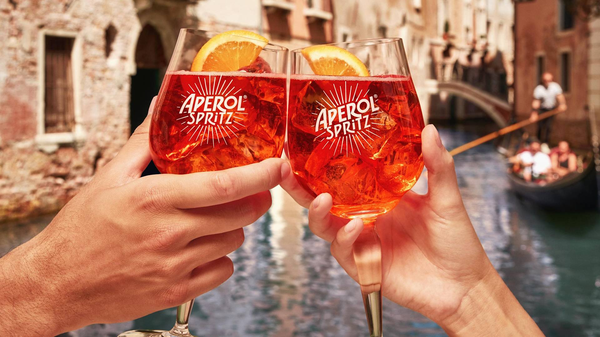 Two hands clink Aperol Spritz glasses garnished with orange slices, with a canal, gondola, and old buildings in the background, suggesting a lively, sunny scene in Venice.