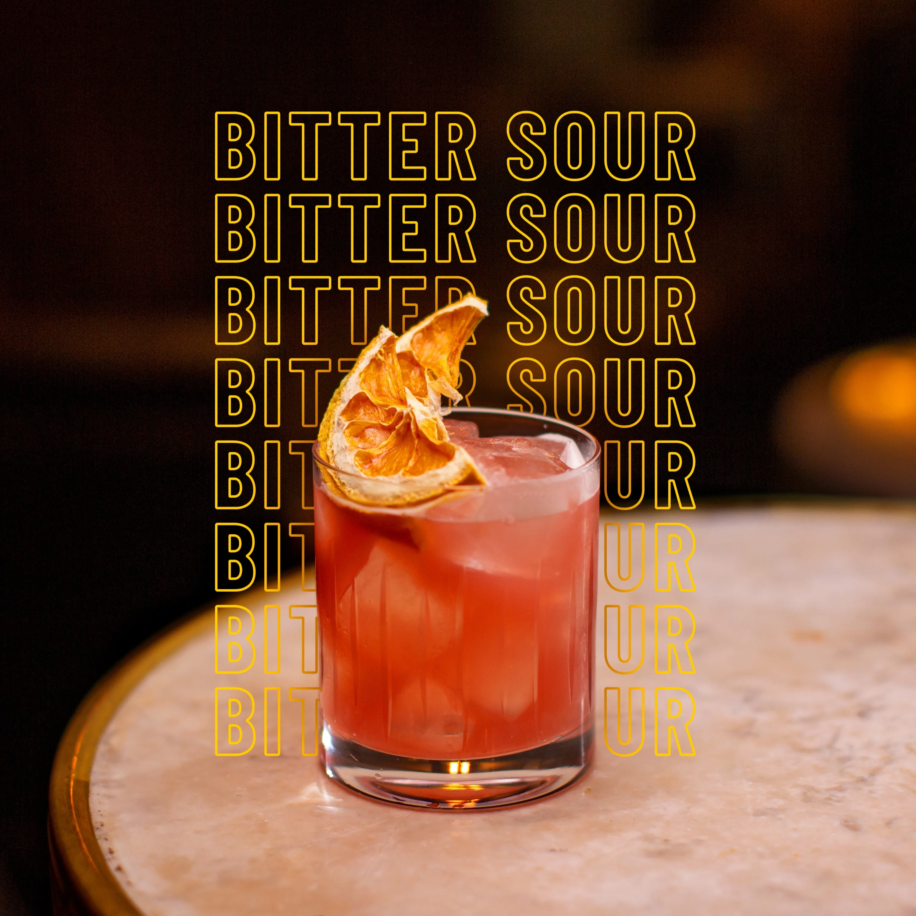 A reddish cocktail garnished with a dried citrus slice sits on a marble table. The words BITTER SOUR are repeated in bold yellow text behind the drink.