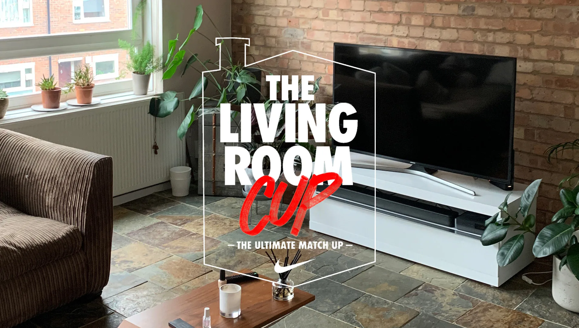 A cozy living room with plants on the windowsill, a brown sofa, a white TV stand with a flat-screen TV, and a coffee table. Text overlay says The Living Room Cup – The Ultimate Match Up.