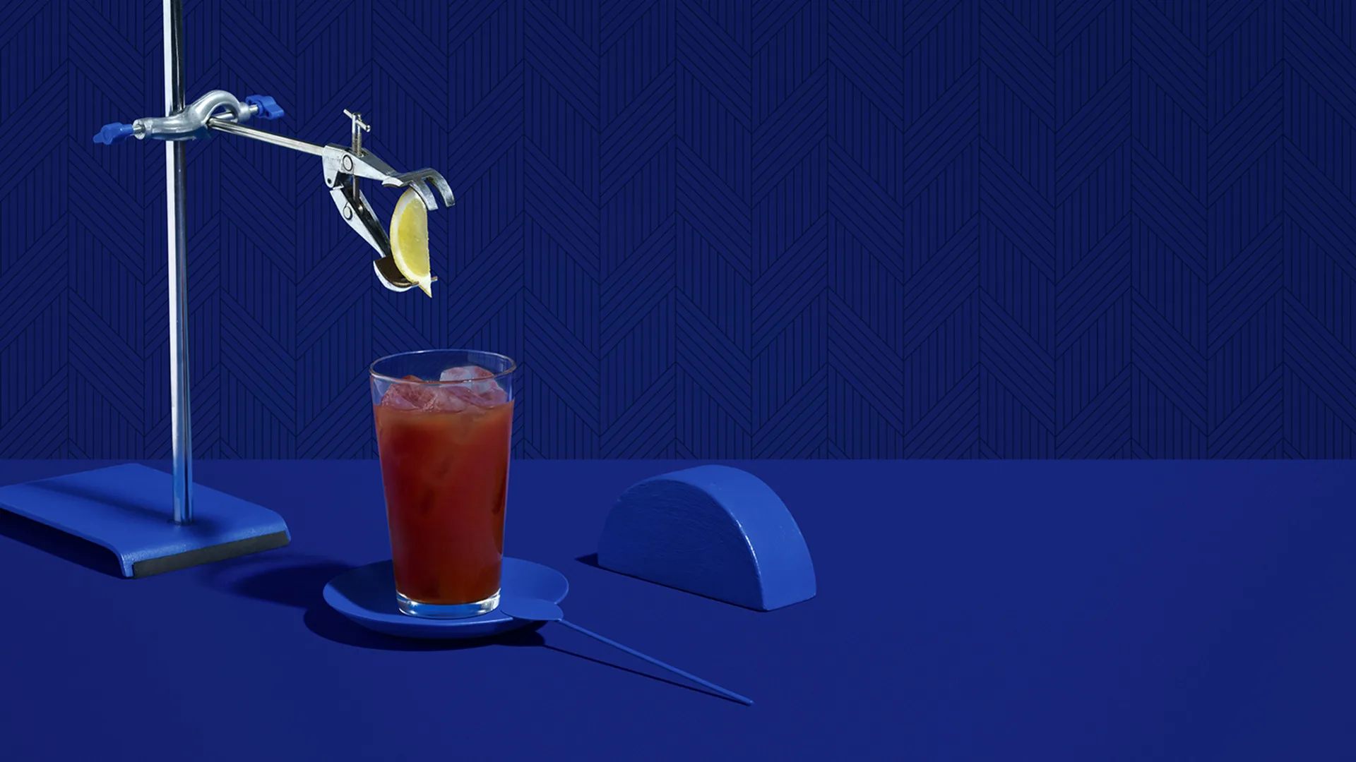 A glass of iced red drink sits on a blue saucer, with a mechanical clamp holding a lime slice above it. The background and table are solid blue, and there is a semicircular blue object beside the glass.