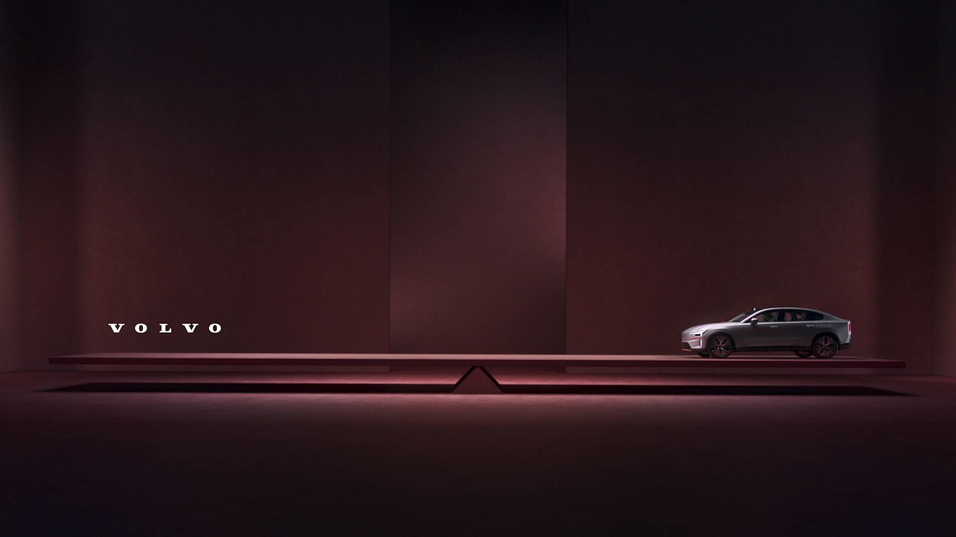 A sleek Volvo sedan is displayed on a raised platform in a minimalistic, dark burgundy room. The Volvo logo is illuminated in white on the left side of the image.