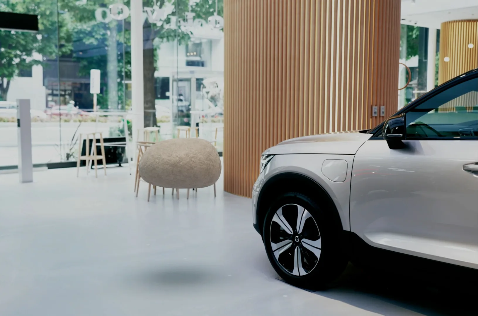 A white electric car is parked indoors next to a large wooden pillar. In the background are glass windows, wooden chairs, and tables. A round beige object sits on the floor near the center of the image.