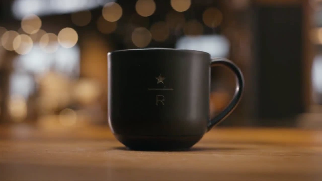 A matte black coffee mug with a handle, featuring a simple star and the letter R design, sits on a wooden surface with blurred, warm lights in the background.