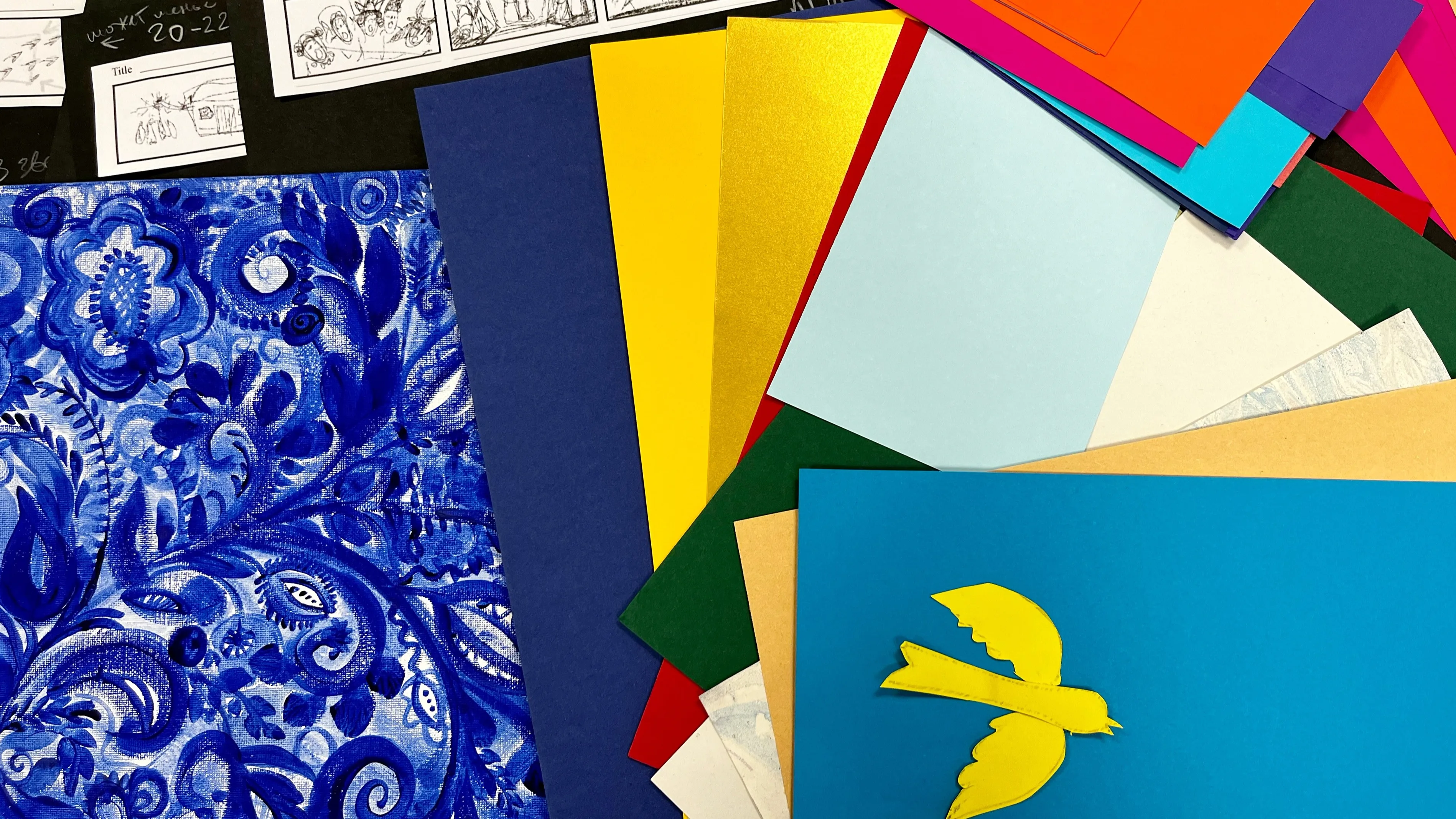 A variety of colorful papers, including blue patterned, red, yellow, and green sheets, are spread out. A yellow paper bird sits on blue paper. Black-and-white comic panels are visible at the top of the image.