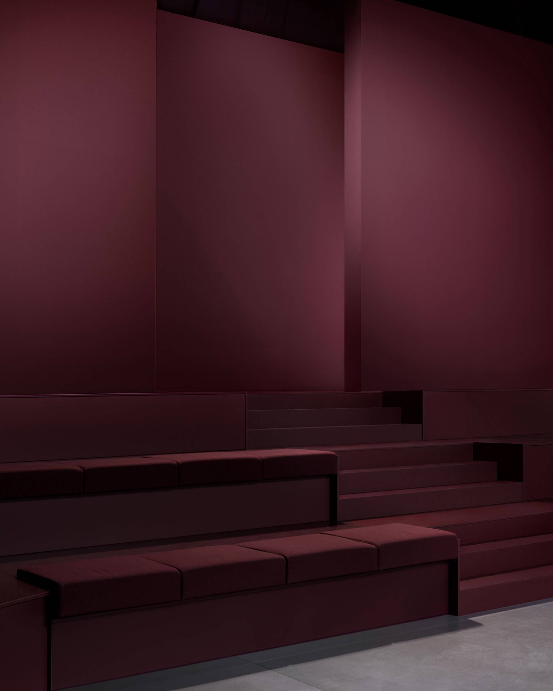 Minimalist interior with tiered maroon seating and matching maroon walls, featuring clean lines and a modern, geometric design. The lighting is soft, creating a calm and sophisticated atmosphere.
