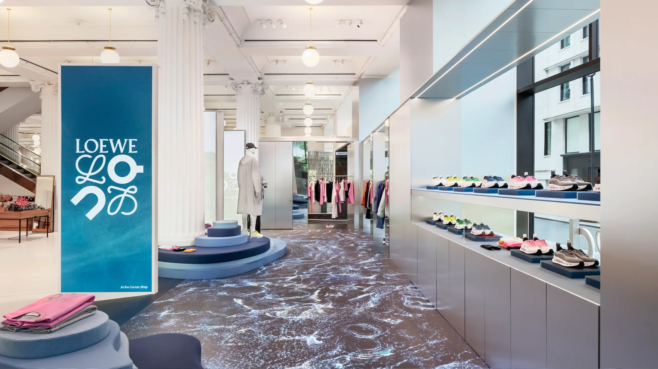 A modern, brightly lit Loewe store with sleek displays of shoes and clothing, white columns, and a large digital Loewe sign. The floor features a swirling, water-like pattern, and large windows let in natural light.