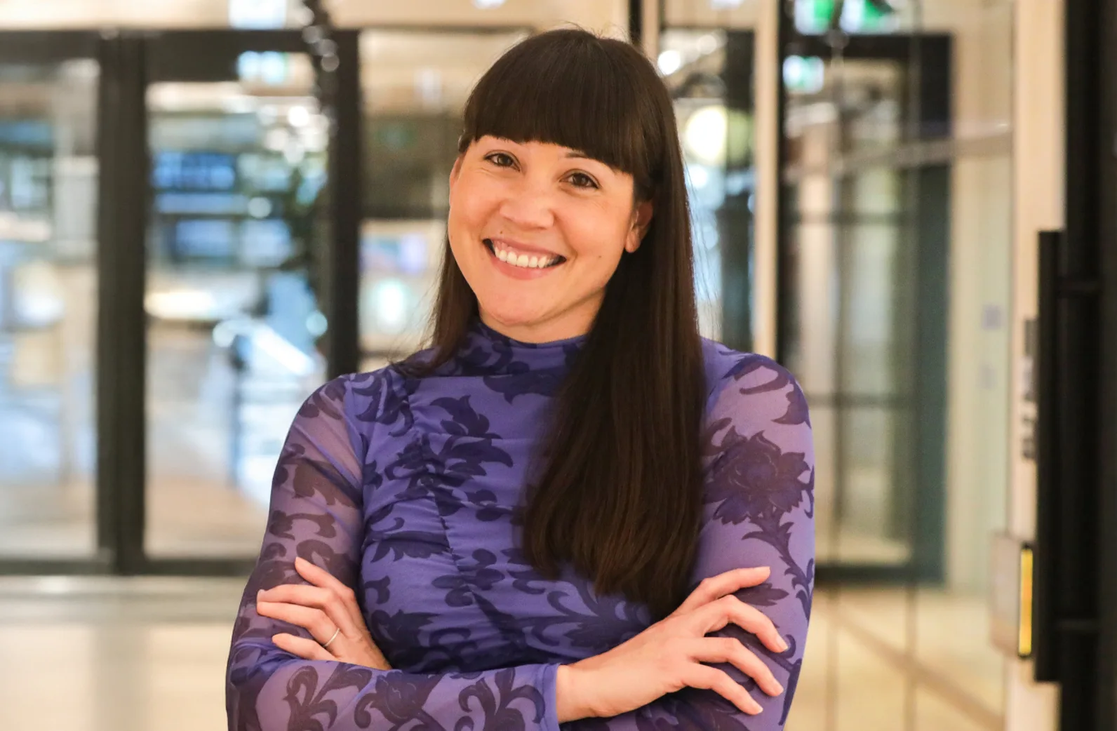 A woman with long dark hair and bangs smiles with arms crossed, wearing a sheer purple floral top, standing indoors with glass doors and a blurred background.
