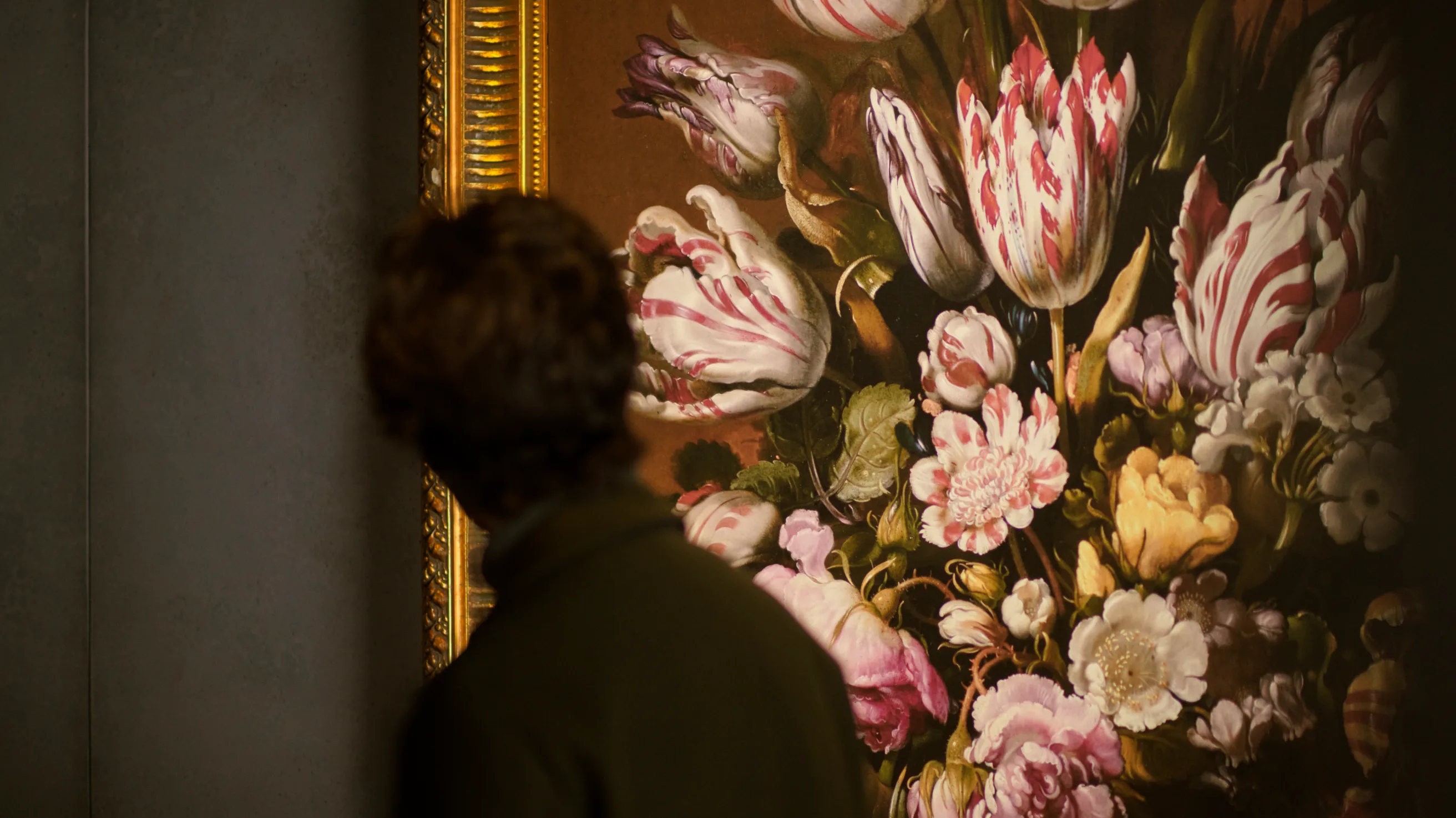 A person with brown hair, seen from behind, closely observes a large framed painting of colorful flowers, including tulips and roses, on a dark background.