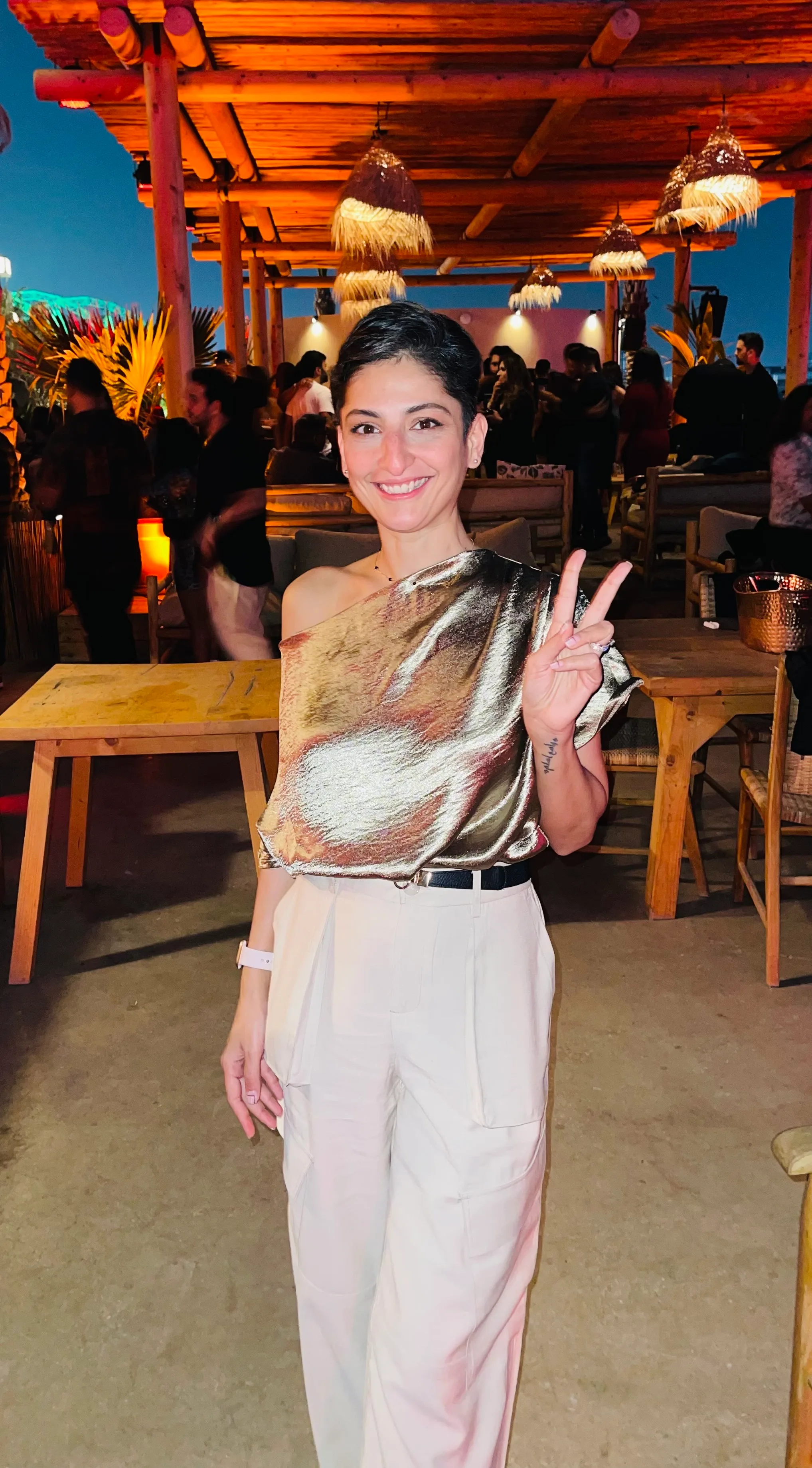 A woman in a shiny, one-shoulder top and light pants stands indoors, smiling and holding up a peace sign. The background shows people, wooden tables, warm lighting, and a casual, lively atmosphere.