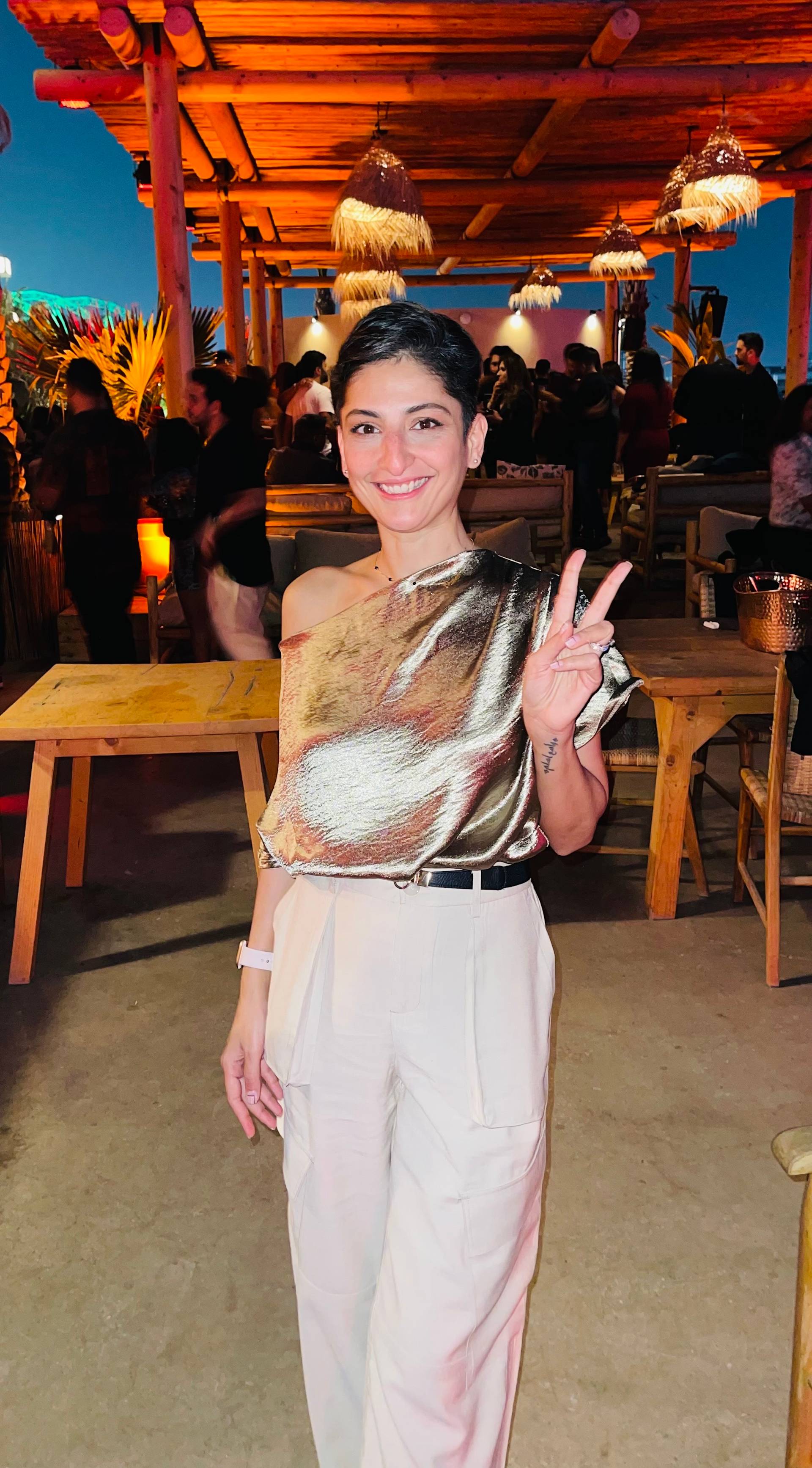 A woman in a shiny, one-shoulder top and light pants stands indoors, smiling and holding up a peace sign. The background shows people, wooden tables, warm lighting, and a casual, lively atmosphere.