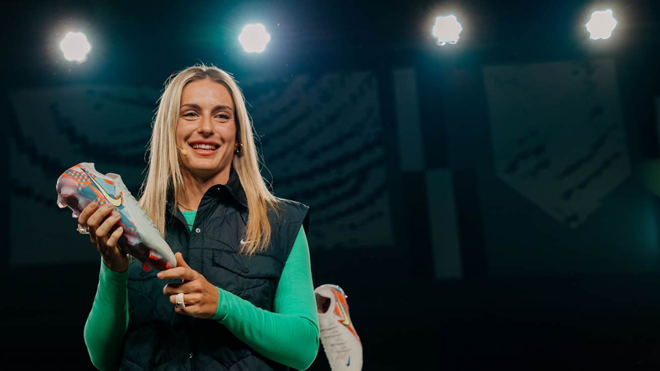 A smiling woman with long blonde hair holds up a colorful athletic shoe on stage under bright lights, wearing a green shirt and dark vest.