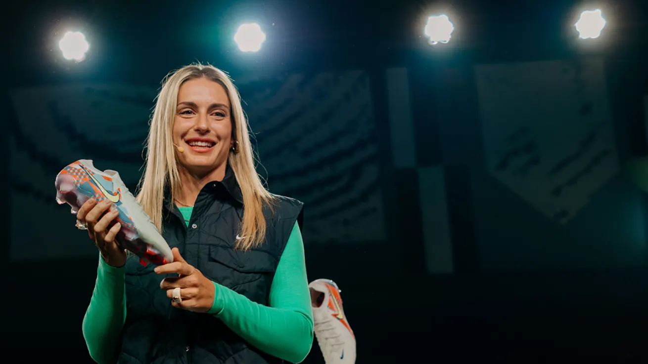 A smiling woman with long blonde hair holds up a colorful athletic shoe on stage under bright lights, wearing a green shirt and dark vest.