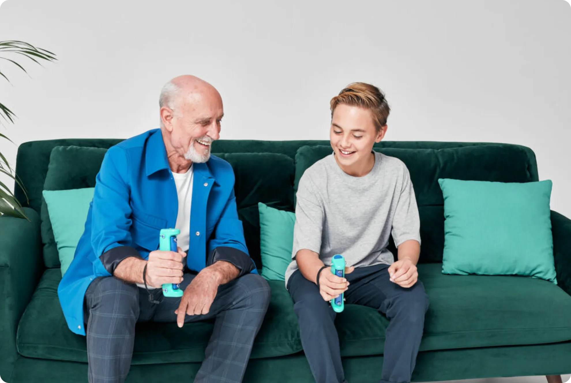 An older man and a boy sit on a green couch, smiling and holding blue game controllers, appearing to play a game together. Four teal pillows are on the couch and a plant is visible in the background.