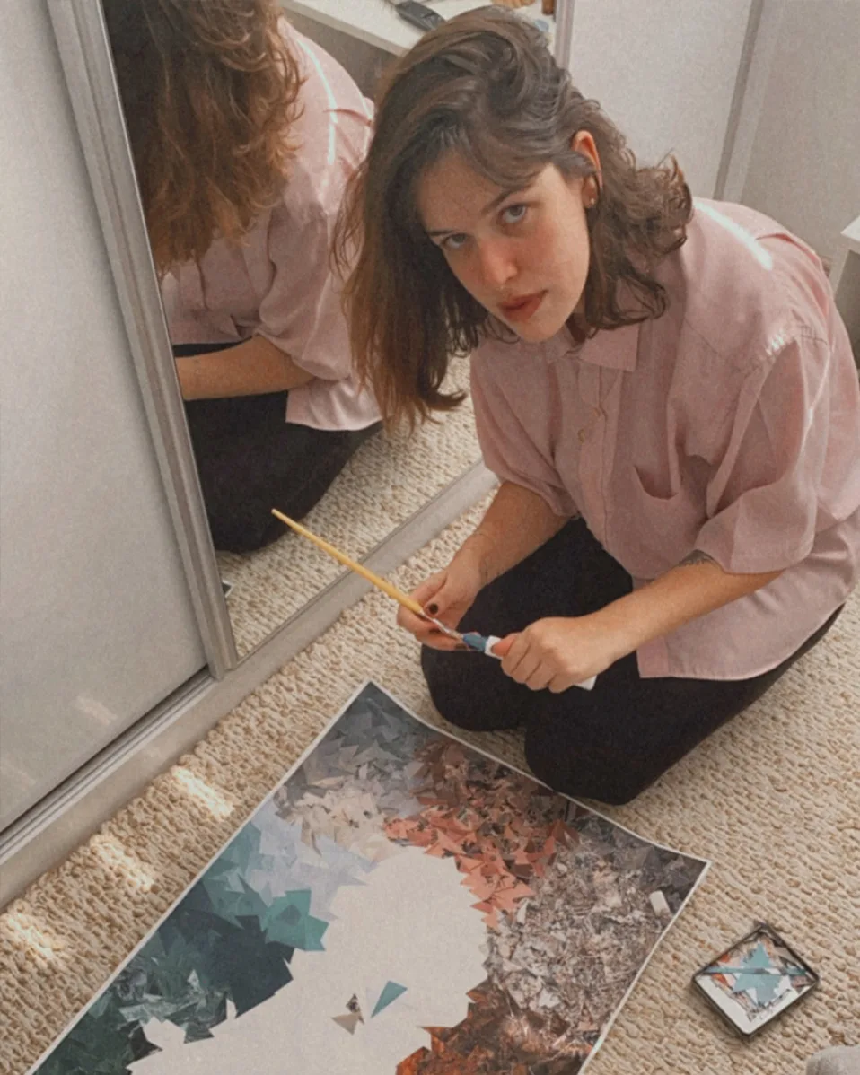 A woman with shoulder-length brown hair wearing a loose pink shirt sits on the floor, holding a paintbrush and working on a colorful abstract painting placed in front of her, with her reflection visible in a mirror beside her.