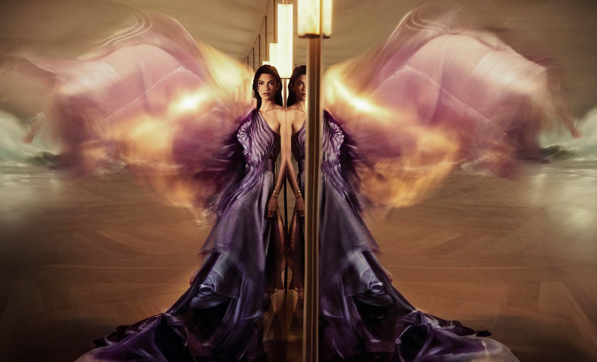 A woman in a flowing, iridescent purple gown stands beside a mirror, creating a symmetrical effect. The fabric fans out dramatically behind her, resembling wings, and the background is softly blurred.
